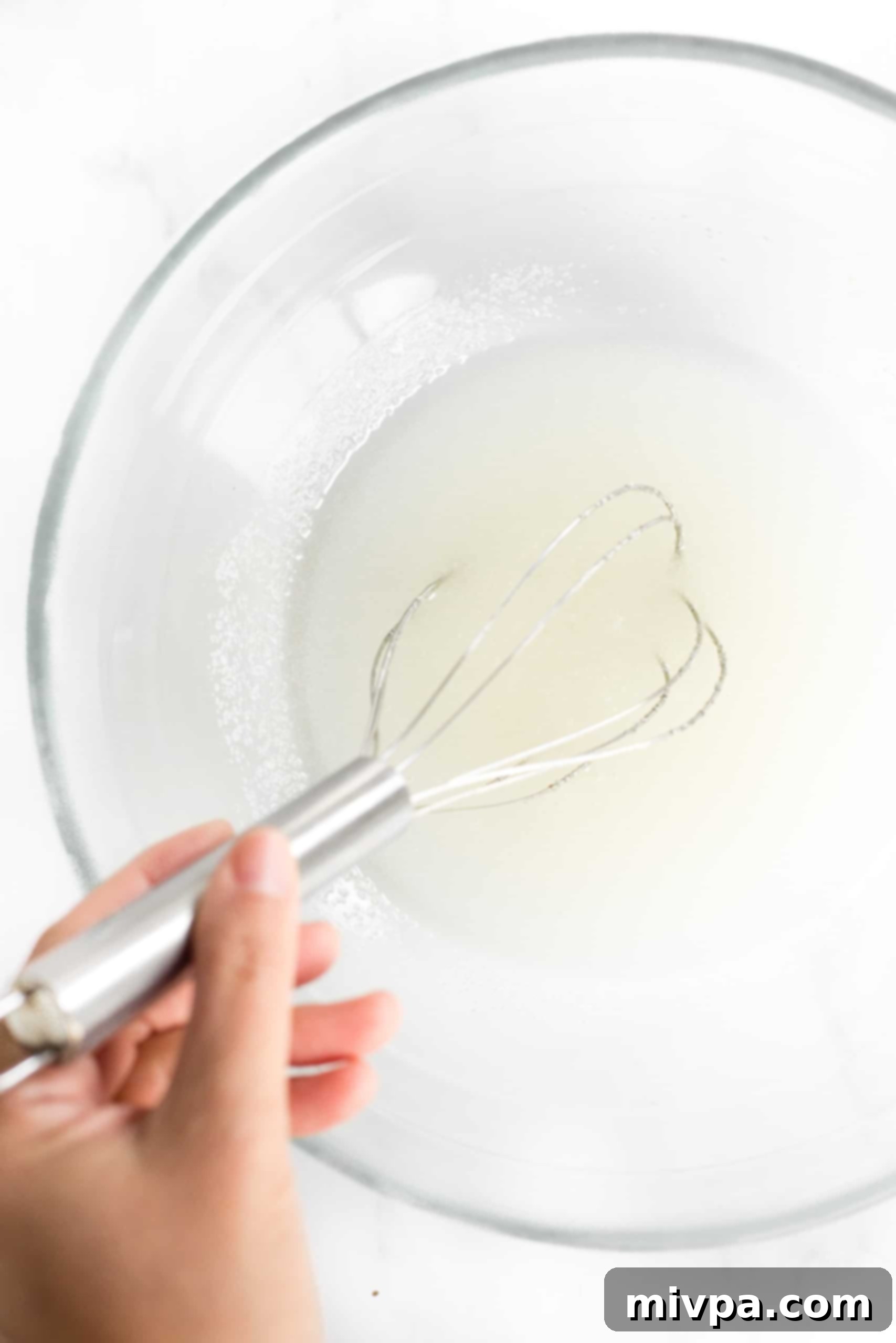A whisk beating oil and sugar together in a large, clear glass bowl.