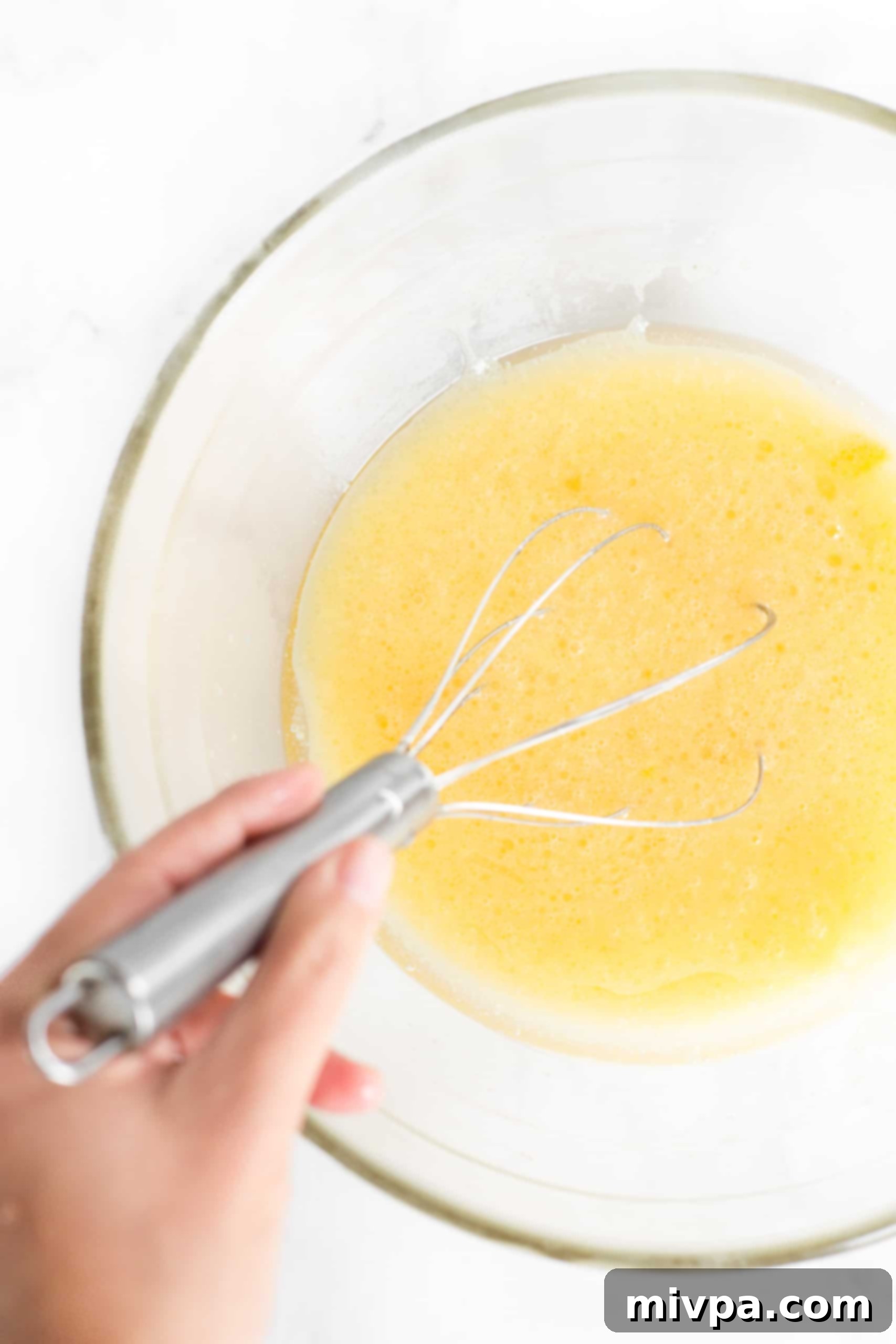A whisk blending a pale yellow liquid mixture in a large glass bowl, containing eggs, milk, sugar, and oil.