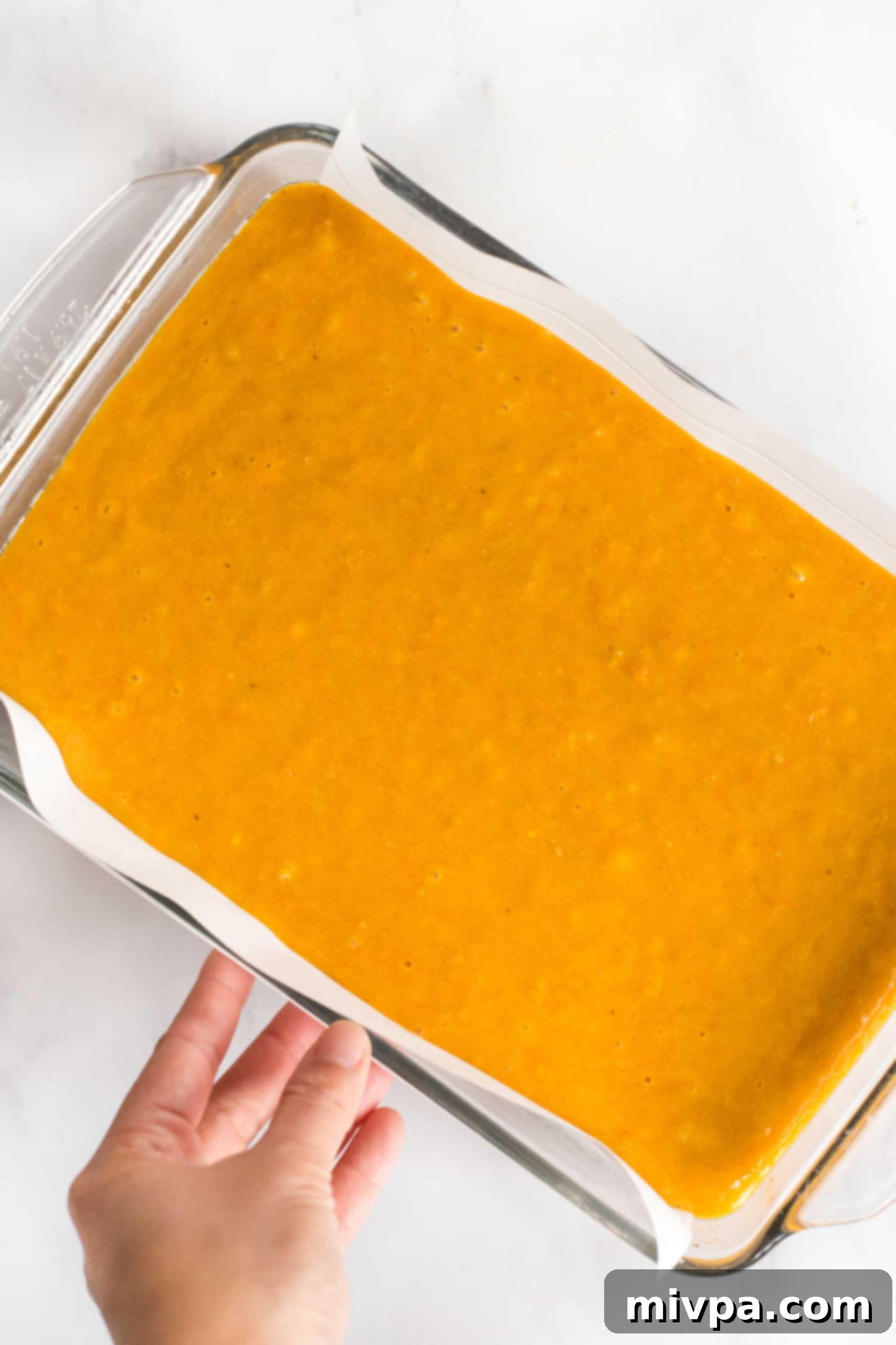 A hand holding a glass baking pan filled with evenly spread pumpkin bar batter, ready for the oven.