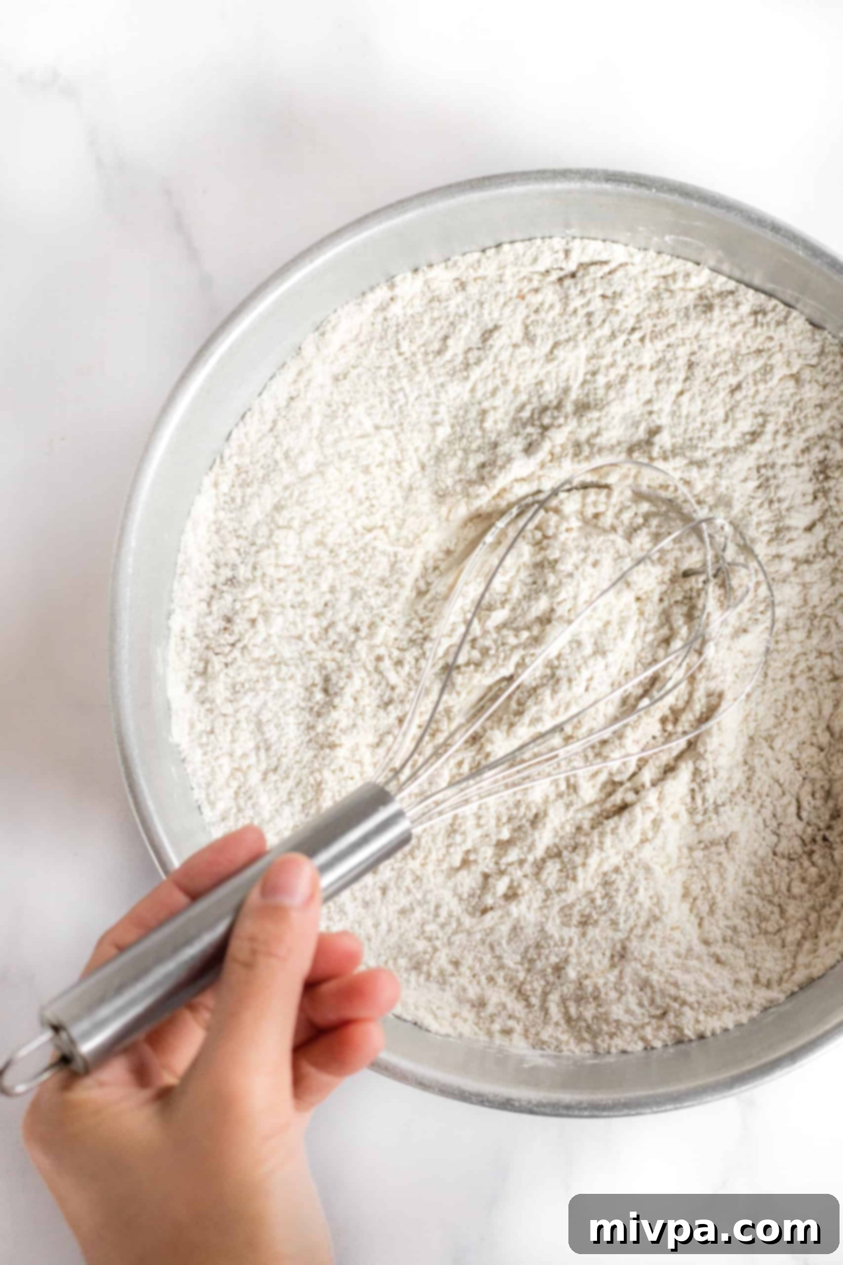 Whisking together the dry ingredients like gluten-free flour, spices, and baking powder in a large metal bowl.