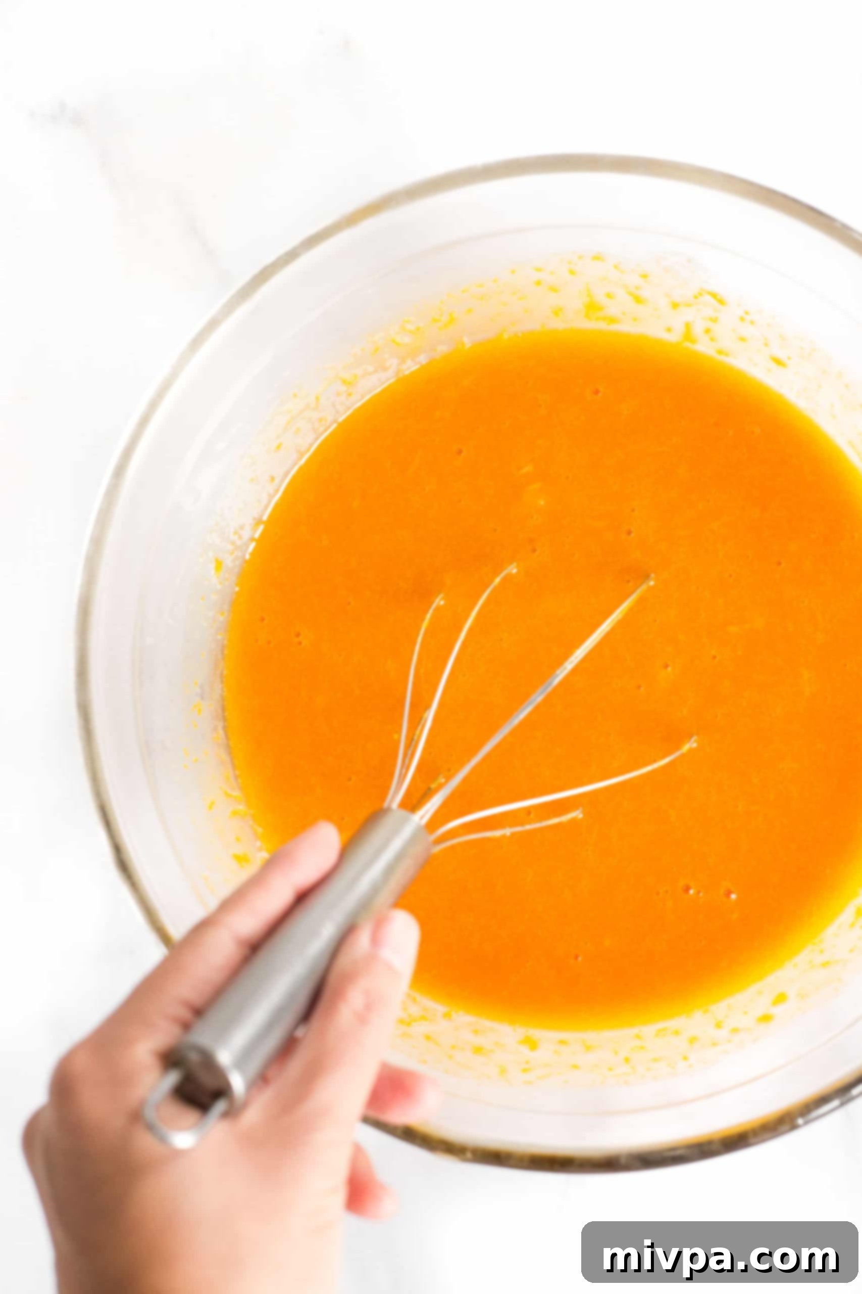 Whisking the bright orange liquid mixture of eggs, vanilla, and pumpkin puree in a glass bowl.