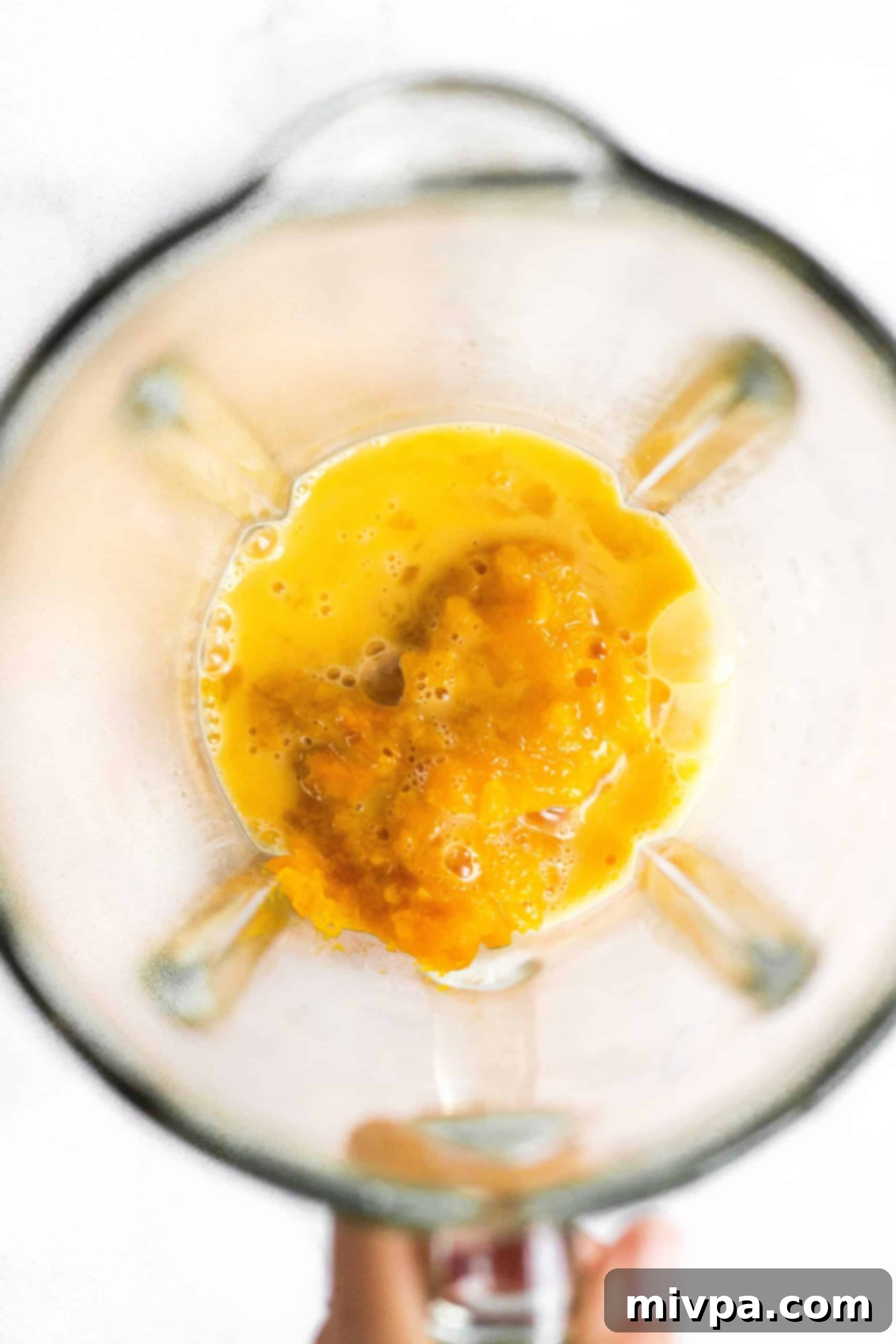A hand holding a blender with a smooth, yellow-orange pumpkin and egg mixture, ready to be incorporated into the bagel dough.
