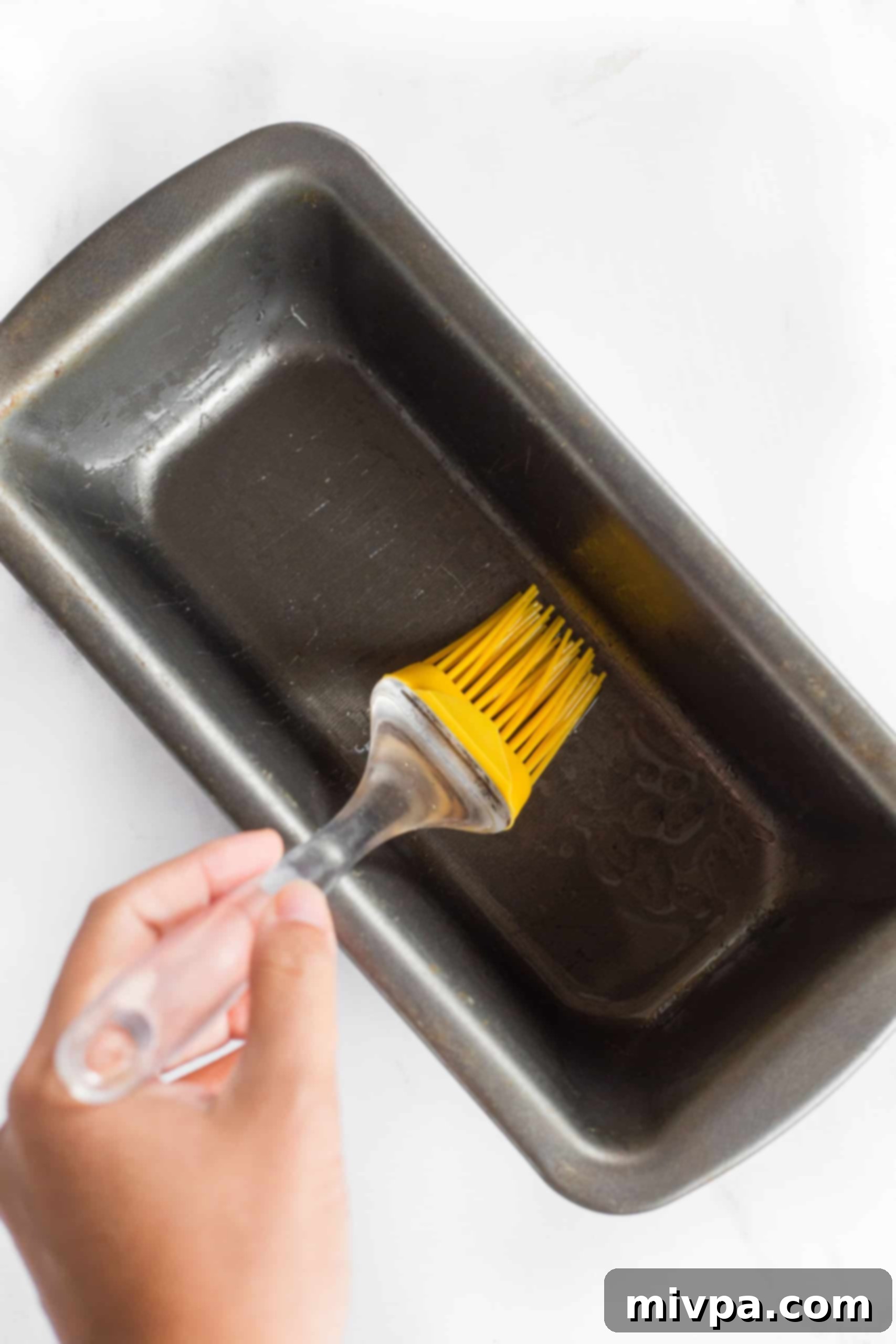 Gluten-Free Whole Grain Bread (Dairy-Free, Vegan) Greasing a metal loaf pan with a yellow silicon brush, preparing it for gluten-free bread dough.