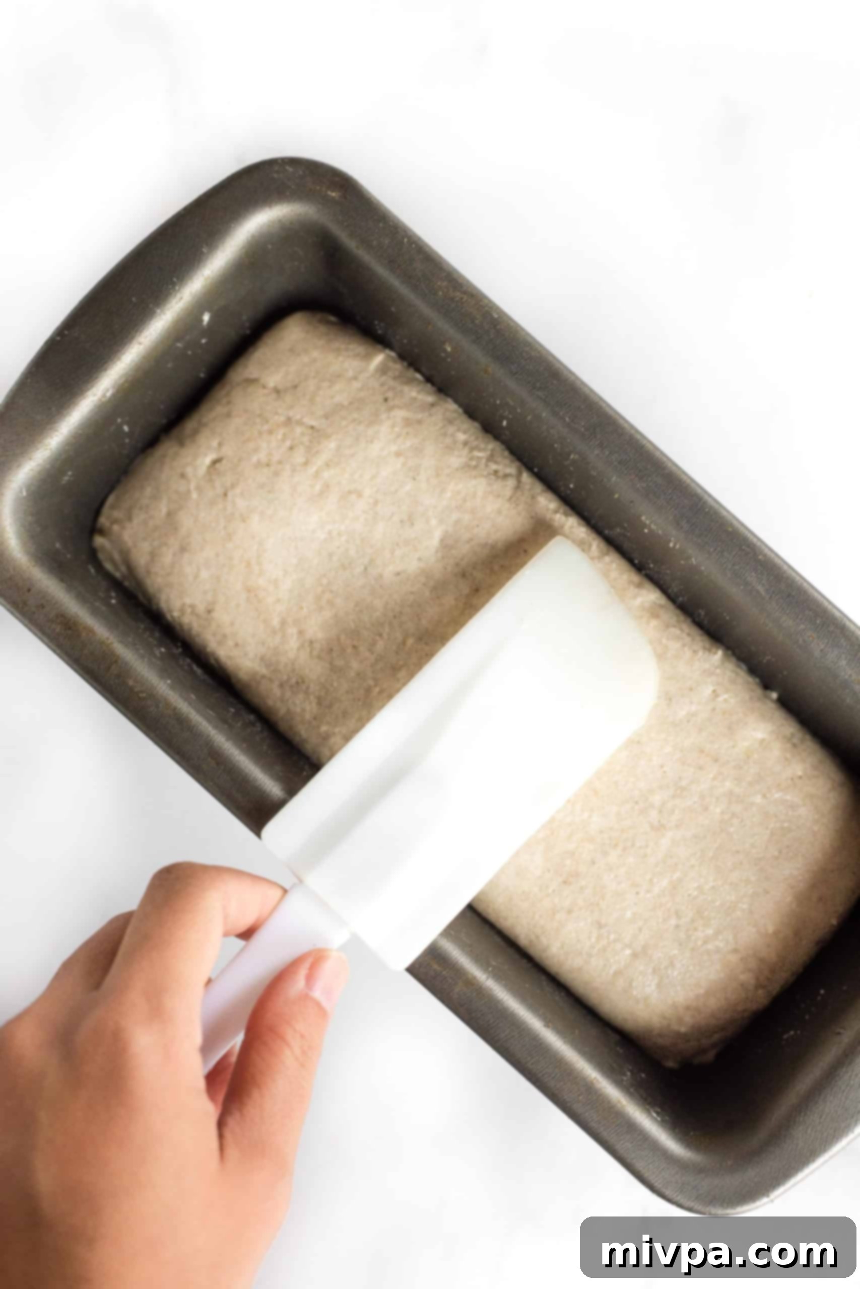 Gluten-Free Whole Grain Bread (Dairy-Free, Vegan) Smoothing out the top of gluten-free brown bread dough in a metal loaf pan with a wet spatula.