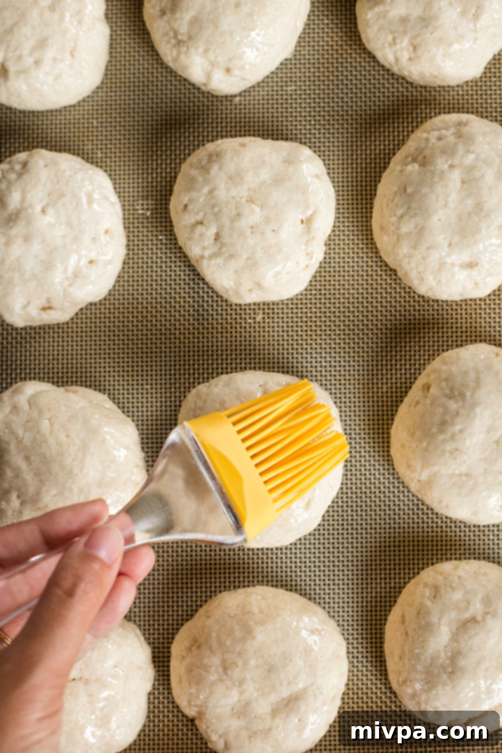 A yellow silicon brush applying oil to risen slider bun dough balls.
