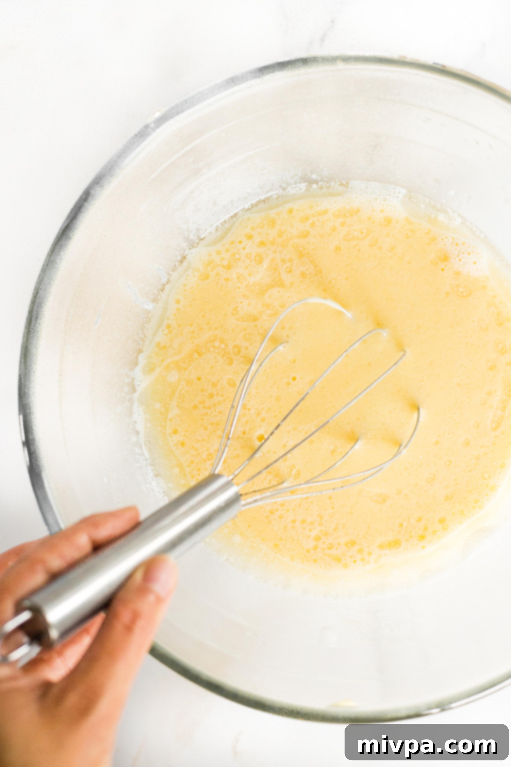 A whisk blending a pale yellow liquid mixture in a large glass mixing bowl.