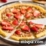 A delicious slice of Chocolate Dessert Pizza, garnished with fresh strawberries and chopped hazelnuts, showing its crispy gluten-free crust and rich ganache.