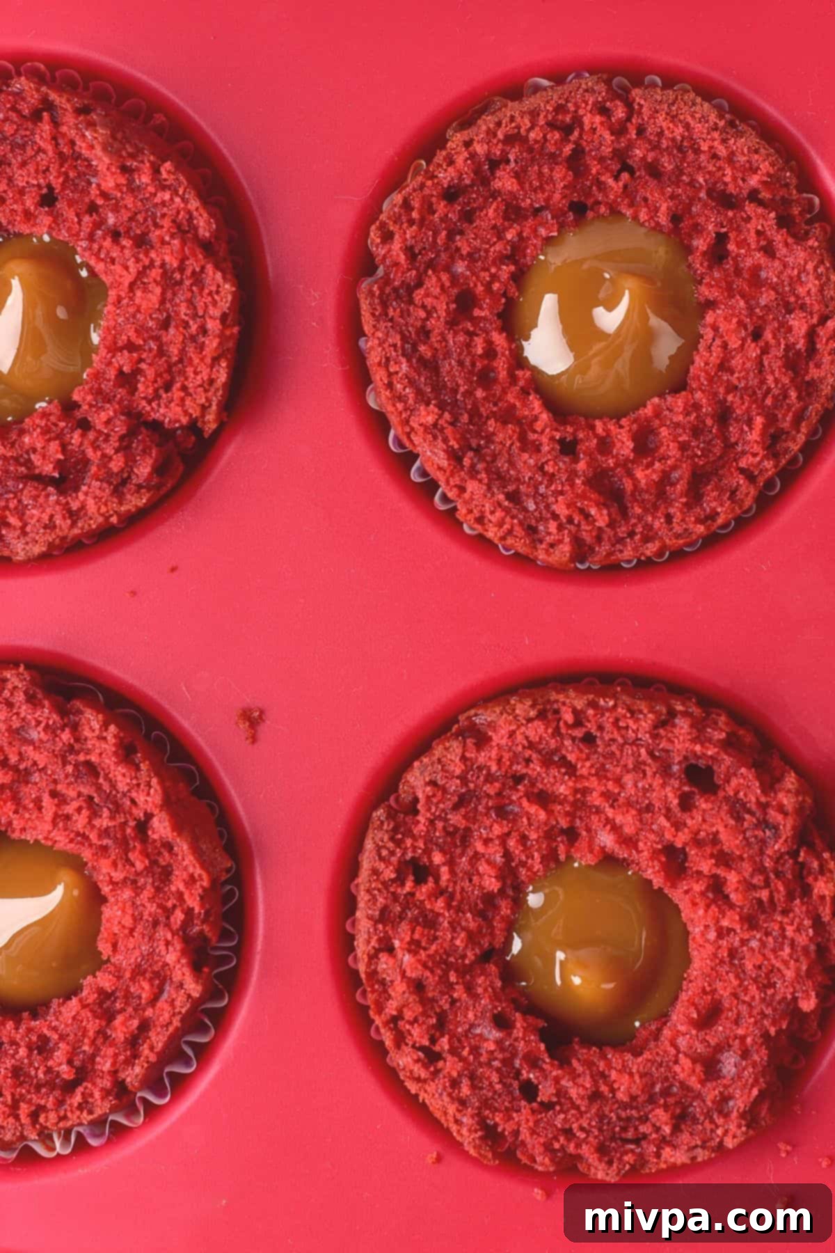 Caramel piped in the middle of red velvet cupcakes in muffin pan.