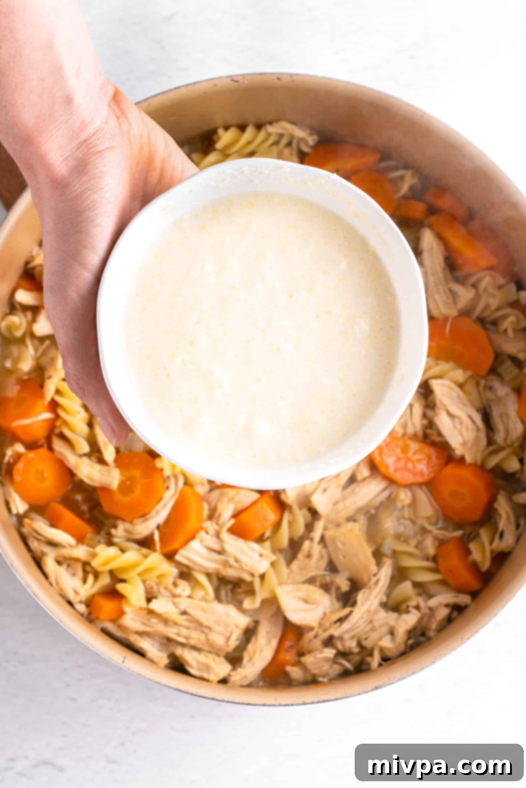 Thickening Gluten-Free Chicken Noodle Soup Recipe (Dairy-Free) with Slurry A hand pouring a bowl of cornstarch slurry, made with cashew milk and dairy-free parmesan, into a pot of gluten-free chicken noodle soup to thicken it.
