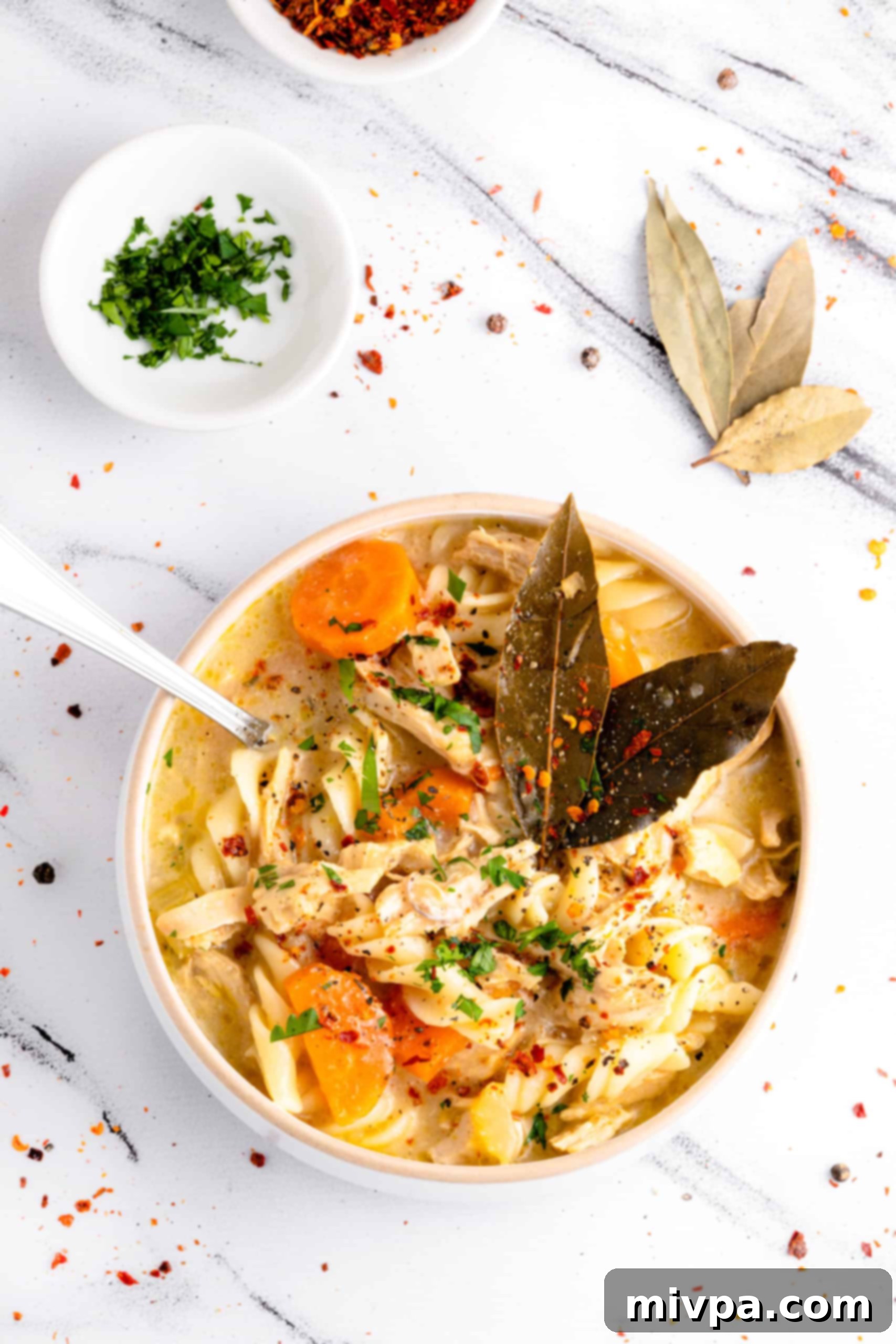 Homemade Gluten-Free Chicken Noodle Soup (Dairy-Free) A top-down view of a hearty bowl of homemade gluten-free chicken noodle soup, garnished with fresh parsley and a sprinkle of chili flakes, served on a marble board.