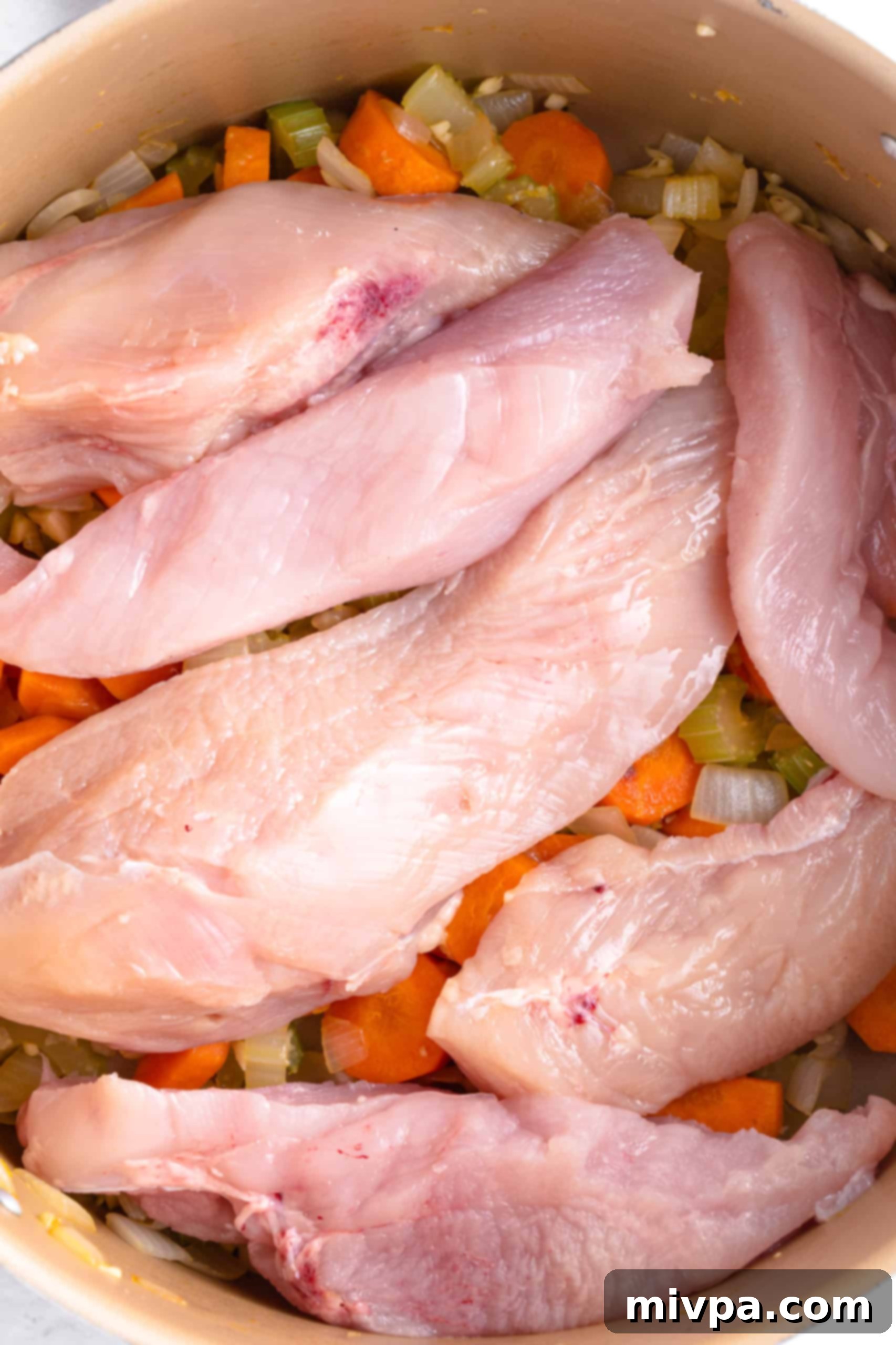 Adding Chicken to Gluten-Free Chicken Noodle Soup Base (Dairy-Free) Raw chicken breast slices added on top of sautéed vegetables in a large pot, ready to be simmered in chicken broth for a flavorful soup.