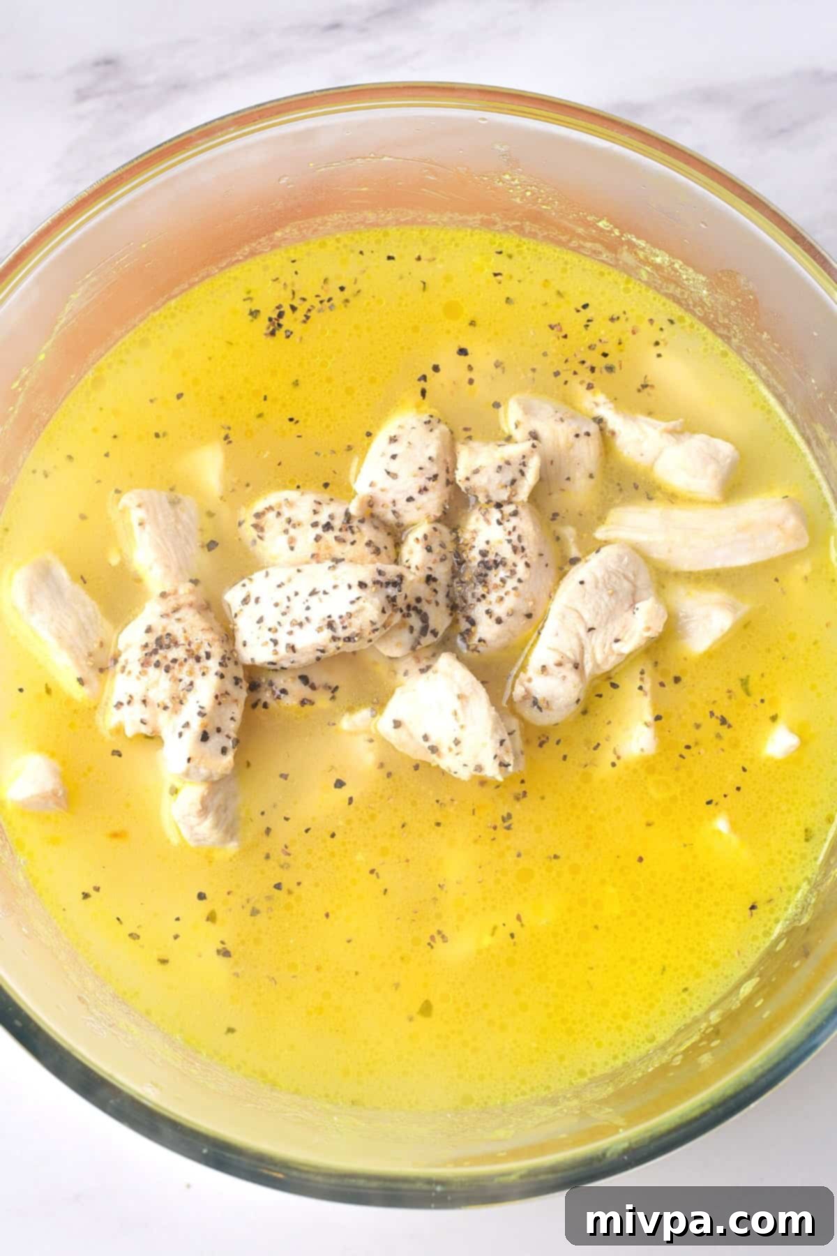 Gluten-Free Chicken Casserole (Dairy-Free) Cooked chicken pieces, rice, onions, and cheese mixed in a yellow liquid within a clear glass bowl, ready for baking.