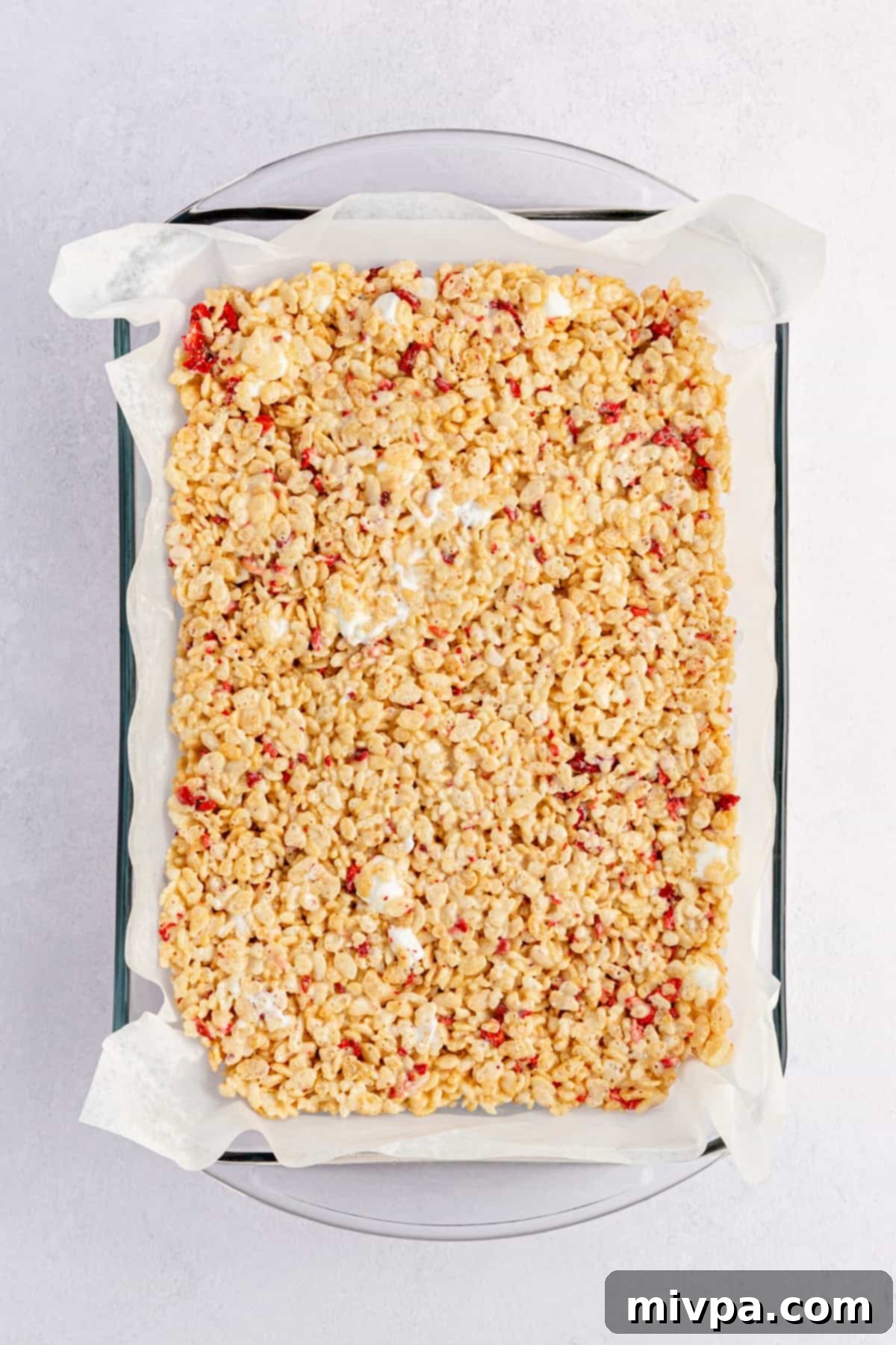 Cereal mixture packed into a parchment-lined baking dish, being flattened with a spatula.