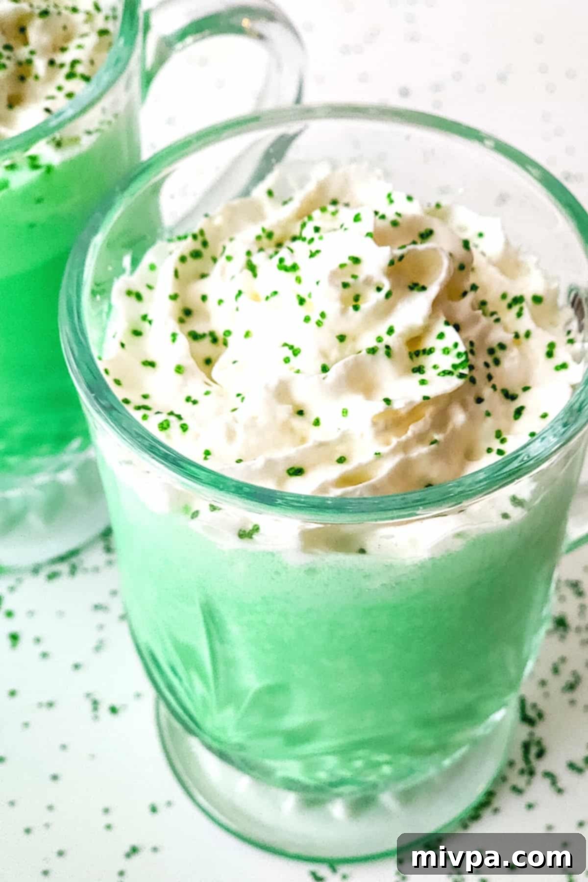 Homemade Vegan Shamrock Shake (Gluten-Free) Two glass cups filled with vibrant green shamrock shakes, topped with fluffy white whipped cream and a sprinkle of festive green sanding sugar.
