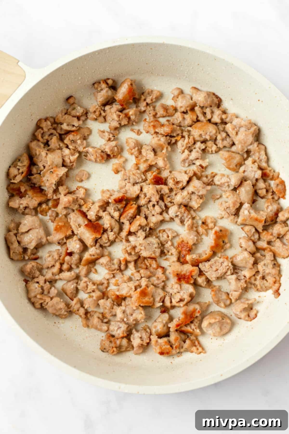 Sausage browning in a skillet, broken into small pieces with a wooden spatula.