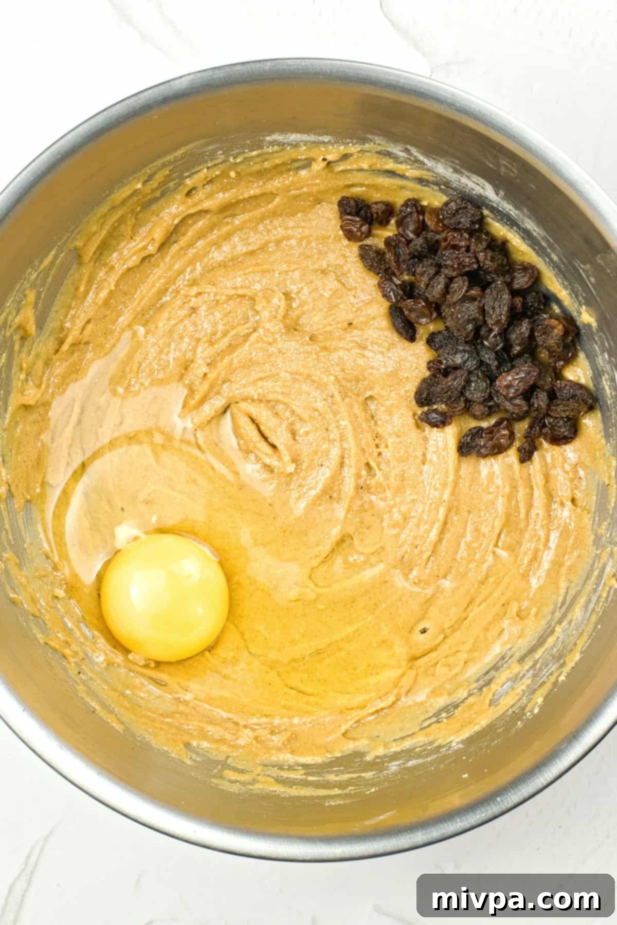 An egg and plump raisins on top of raw gluten-free bread dough in a metal bowl.