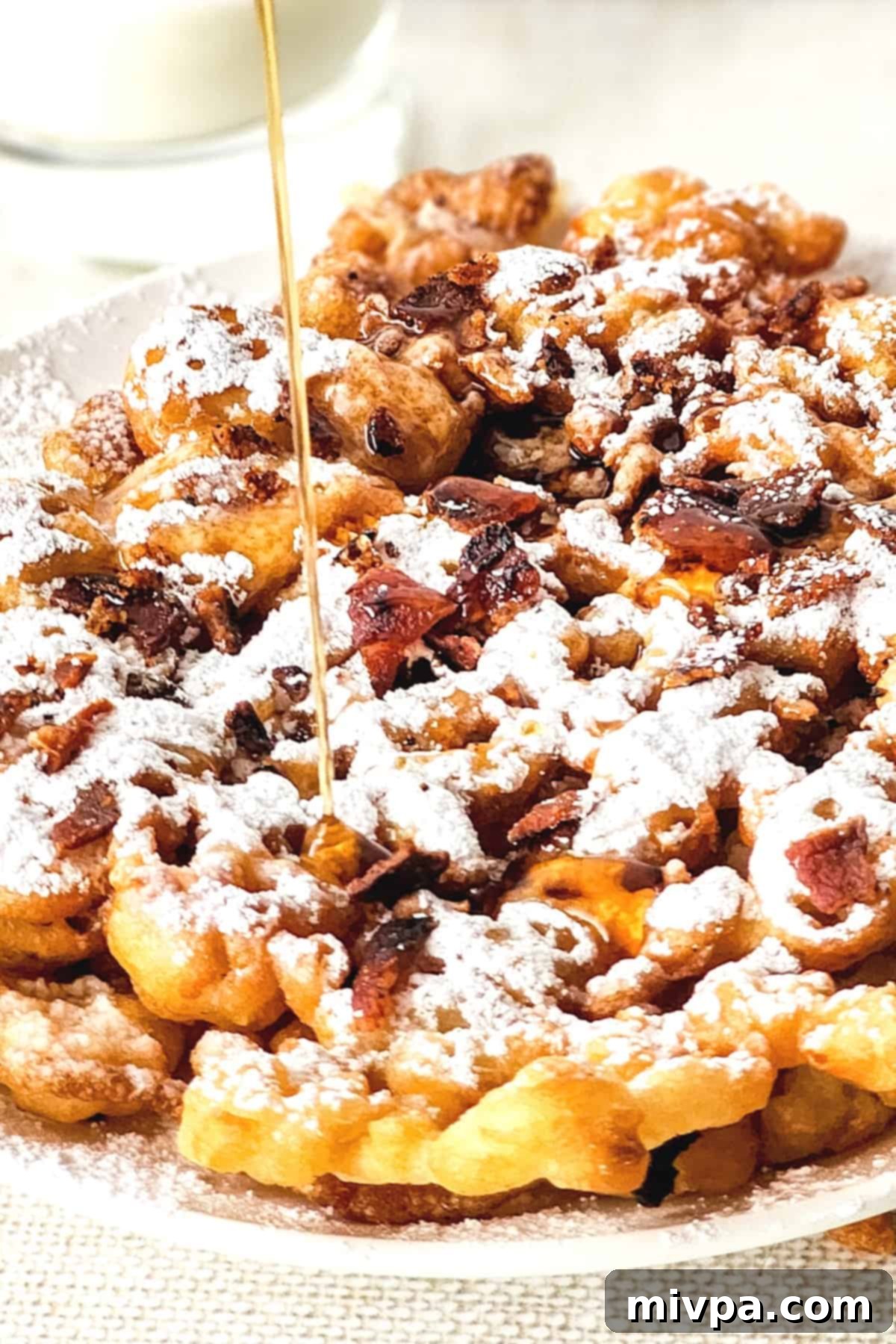 A hand drizzling maple syrup over a stack of warm gluten-free funnel cakes.