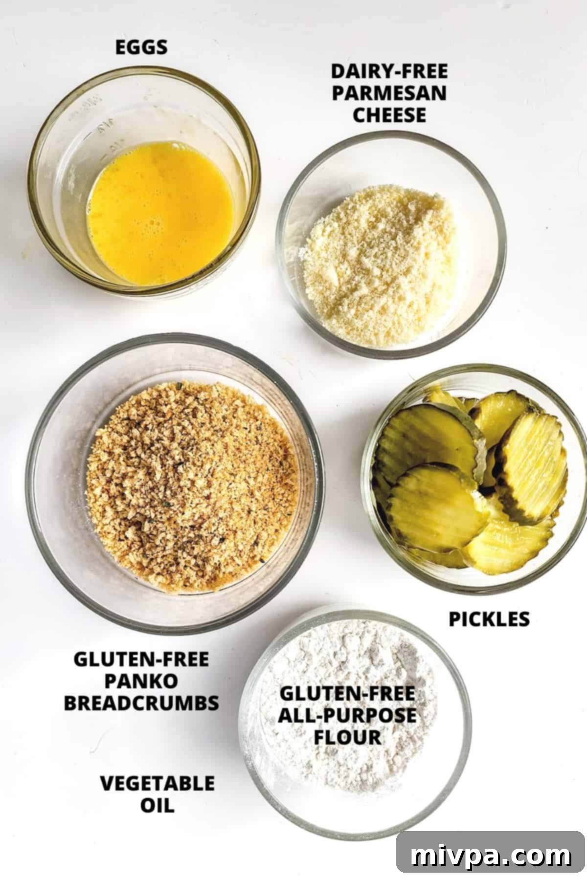 Ingredients for gluten-free fried pickles recipe including gluten-free panko, dairy-free parmesan, gluten-free flour, eggs, and pickle slices, neatly laid out on a white board.