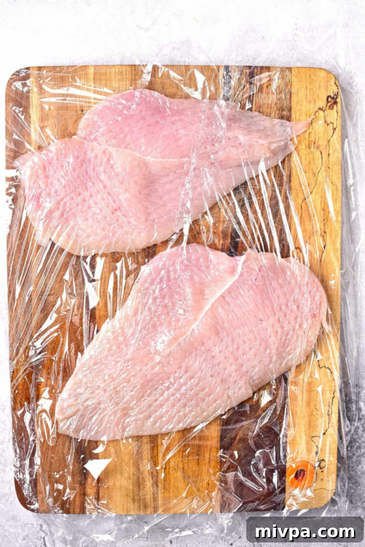 Chicken cutlets on wooden board covered with plastic wrap.