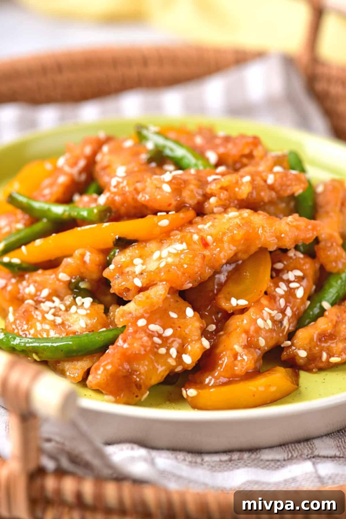 A vibrant plate of homemade honey sesame chicken, featuring crispy chicken pieces, bright yellow bell peppers, and fresh green beans, all generously coated in a glossy, sweet-and-sour sauce and sprinkled with sesame seeds. The dish is presented on a rustic wooden surface, highlighting its appetizing appeal. This healthier take on the popular American Chinese takeout favorite is also gluten-free and dairy-free, making it perfect for a delicious family dinner.