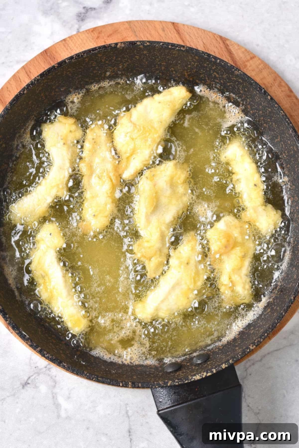 Golden-brown, flour-coated chicken strips are sizzling and frying in hot oil in a medium skillet. The oil bubbles around the chicken, indicating it's at the perfect temperature for achieving a crispy texture. This action shot captures the crucial stage of cooking the chicken to perfection.