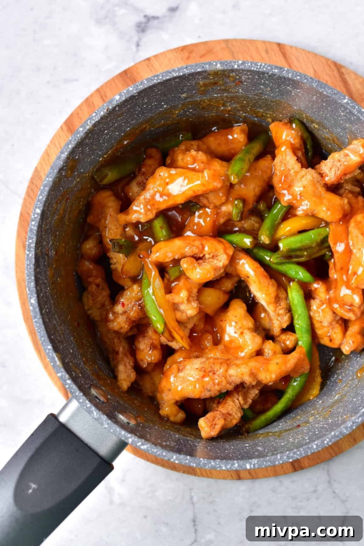 Crispy fried chicken strips are being tossed in a rich, glossy honey sesame sauce within a saucepan. The chicken pieces are evenly coated, reflecting the light and showing the perfect texture before serving. This step captures the final delicious combination of ingredients.