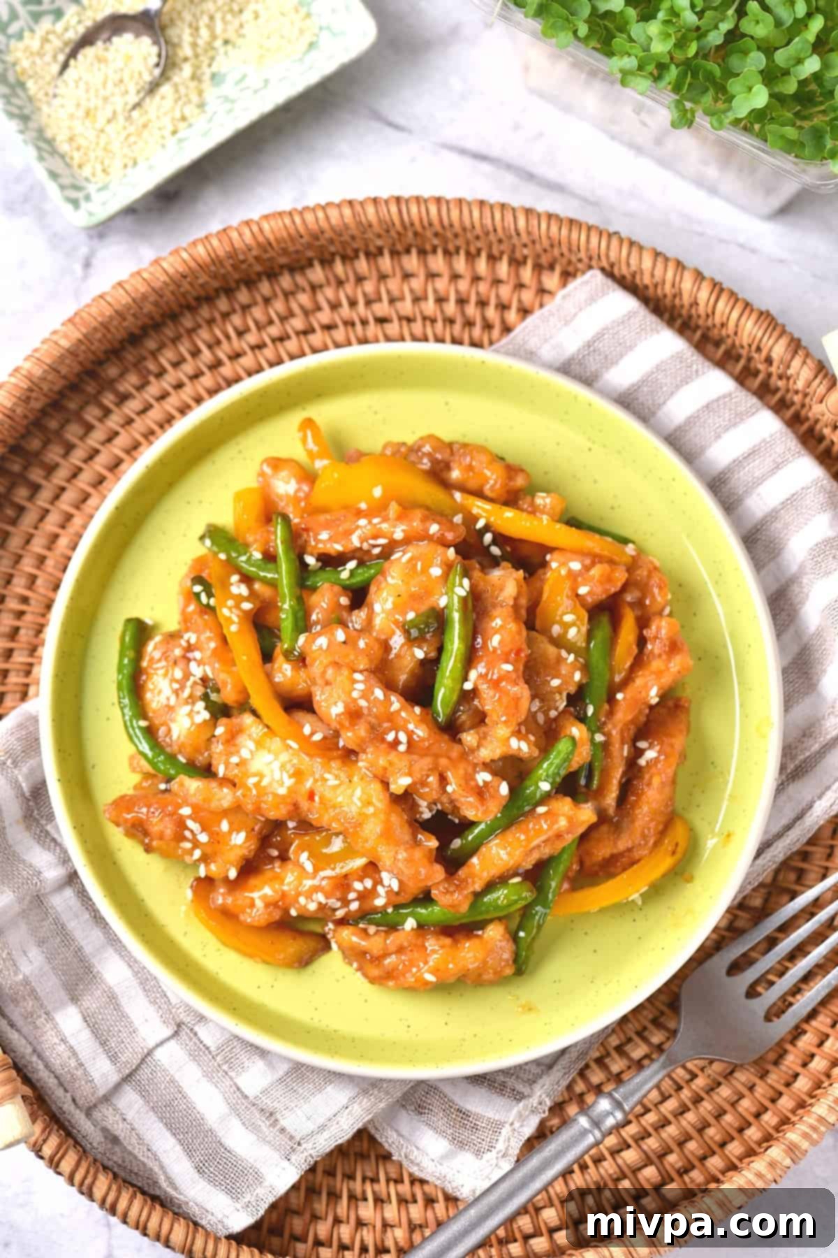 A top-down view of a beautifully plated honey sesame chicken dish resting on a patterned kitchen towel. The crispy chicken pieces, mixed with vibrant bell peppers and green beans, are coated in a glossy, golden sauce and sprinkled generously with white sesame seeds. This enticing presentation highlights the freshness and homemade quality of the Panda Express copycat recipe.