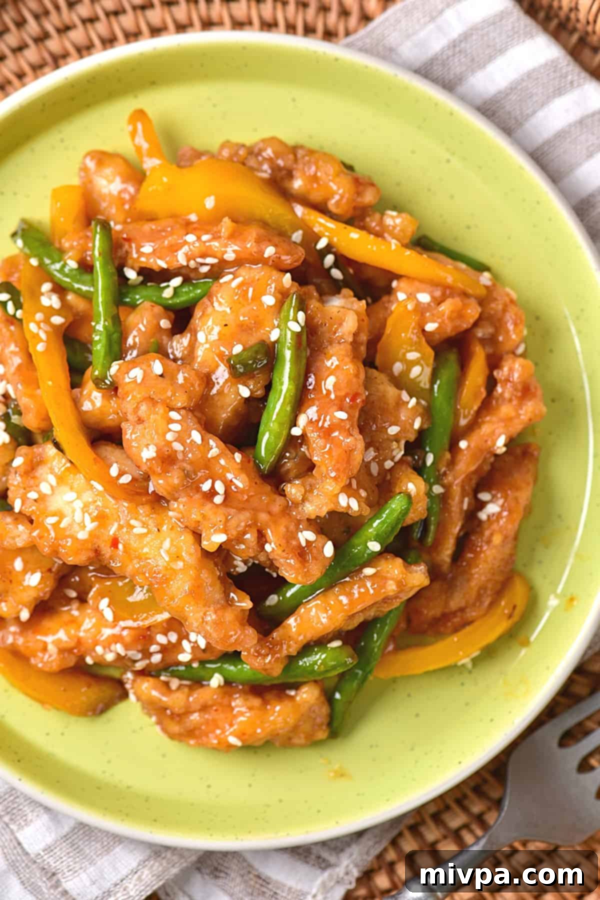 A close-up shot of a plate brimming with golden-brown honey sesame chicken, glistening with its sweet and tangy sauce, accompanied by tender-crisp bell peppers and green beans. White sesame seeds are sprinkled generously over the dish, enhancing its visual appeal and promising a delightful crunch. The focus highlights the delicious texture and vibrant colors of this homemade Panda Express copycat.