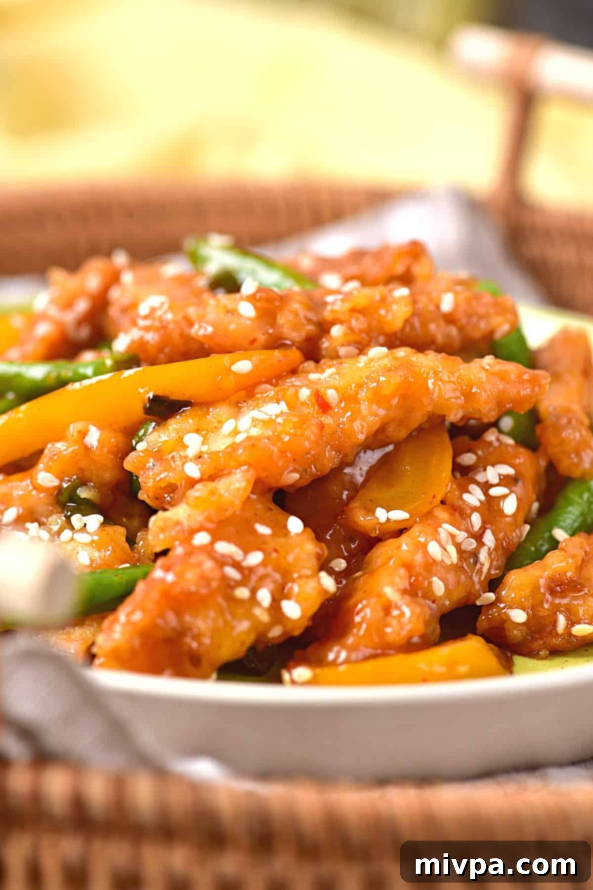 A beautifully arranged plate of gluten-free Panda Express copycat sesame chicken sits invitingly in a rustic wicker basket on a light-colored surface. The crispy chicken pieces, coated in a rich honey sesame sauce, are mixed with bright yellow bell peppers and green beans, all sprinkled with sesame seeds. This image emphasizes the homemade, wholesome, and delicious nature of the gluten-free and dairy-free dish.