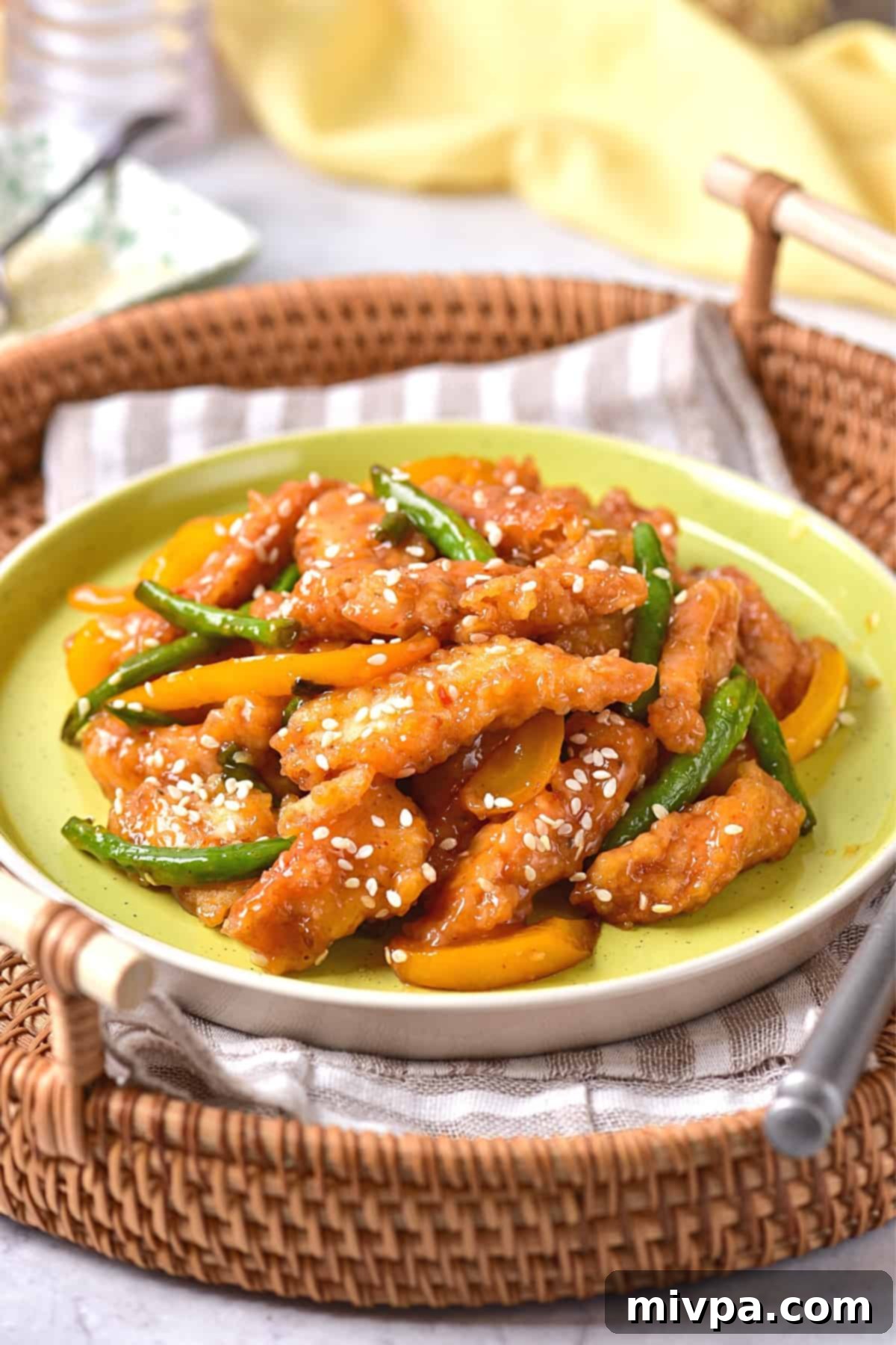 A close-up, top-down view of a serving of homemade honey sesame chicken, presented in a bowl within a larger wicker basket. The crispy chicken pieces are coated in a rich, glossy sauce, vibrant with yellow bell peppers and green beans, and garnished with white sesame seeds. The focus on the dish highlights its appealing textures and colors, emphasizing a healthy, gluten-free, and dairy-free meal option.