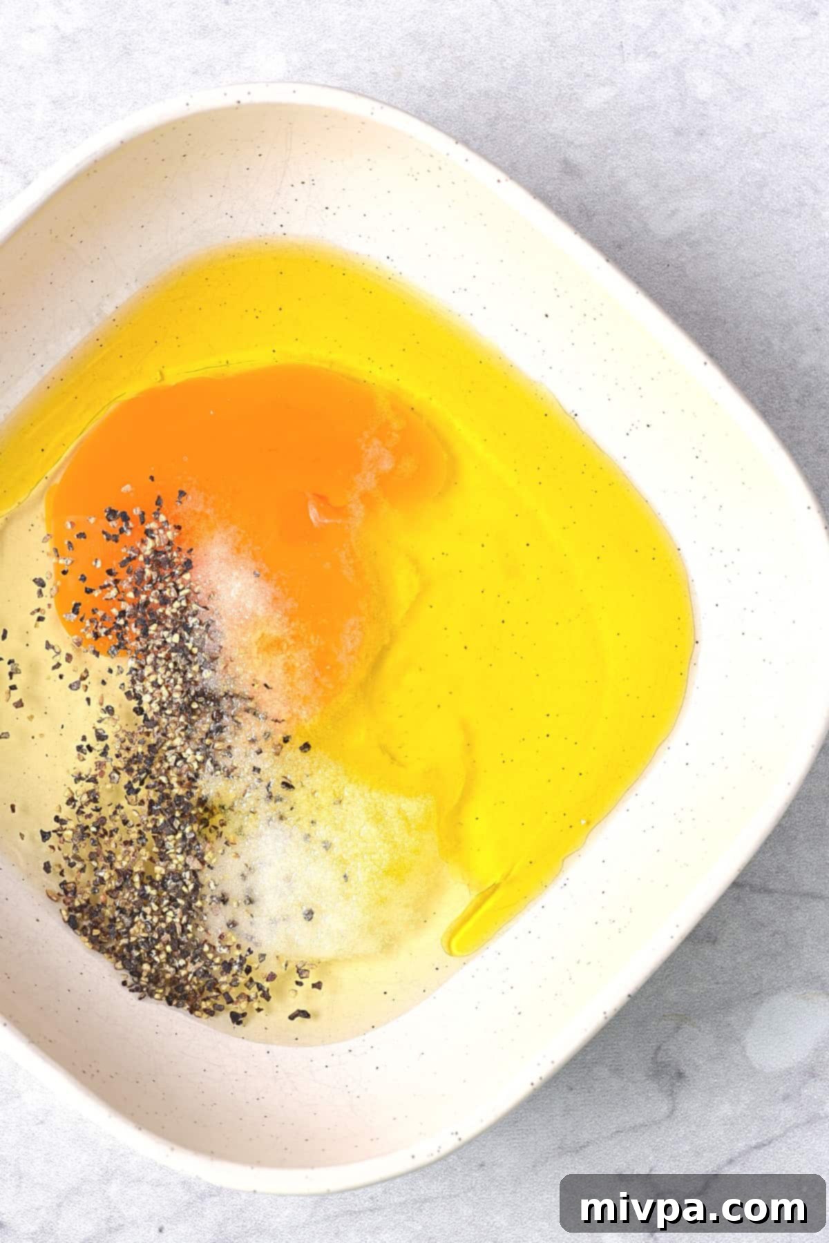 A white bowl containing an egg, olive oil, salt, and pepper, all beaten together. The ingredients are perfectly combined, creating a smooth, pale yellow mixture, ready for coating the chicken. This image emphasizes the initial preparation step for the gluten-free honey sesame chicken recipe.