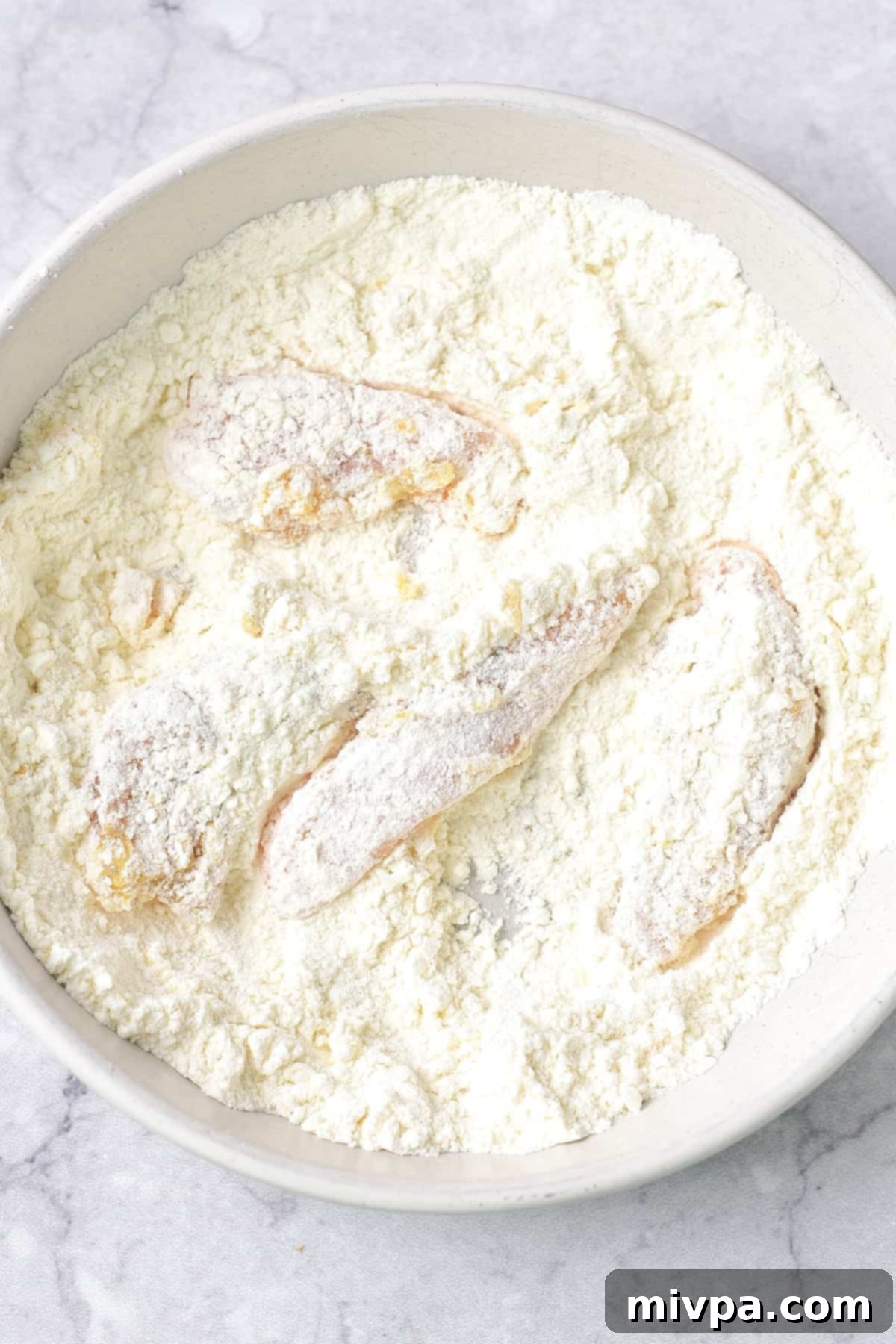 Chicken strips are being carefully rolled and coated in the gluten-free flour mixture, ensuring an even and thorough covering. Excess flour is gently shaken off each strip, highlighting the delicate balance required for a perfectly crispy texture once fried.