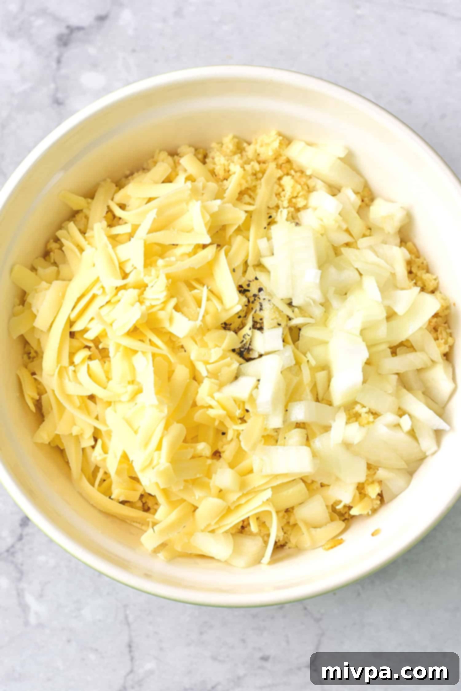 Shredded cheese and diced onions on top of processed hash browns in a bowl, ready to be mixed.