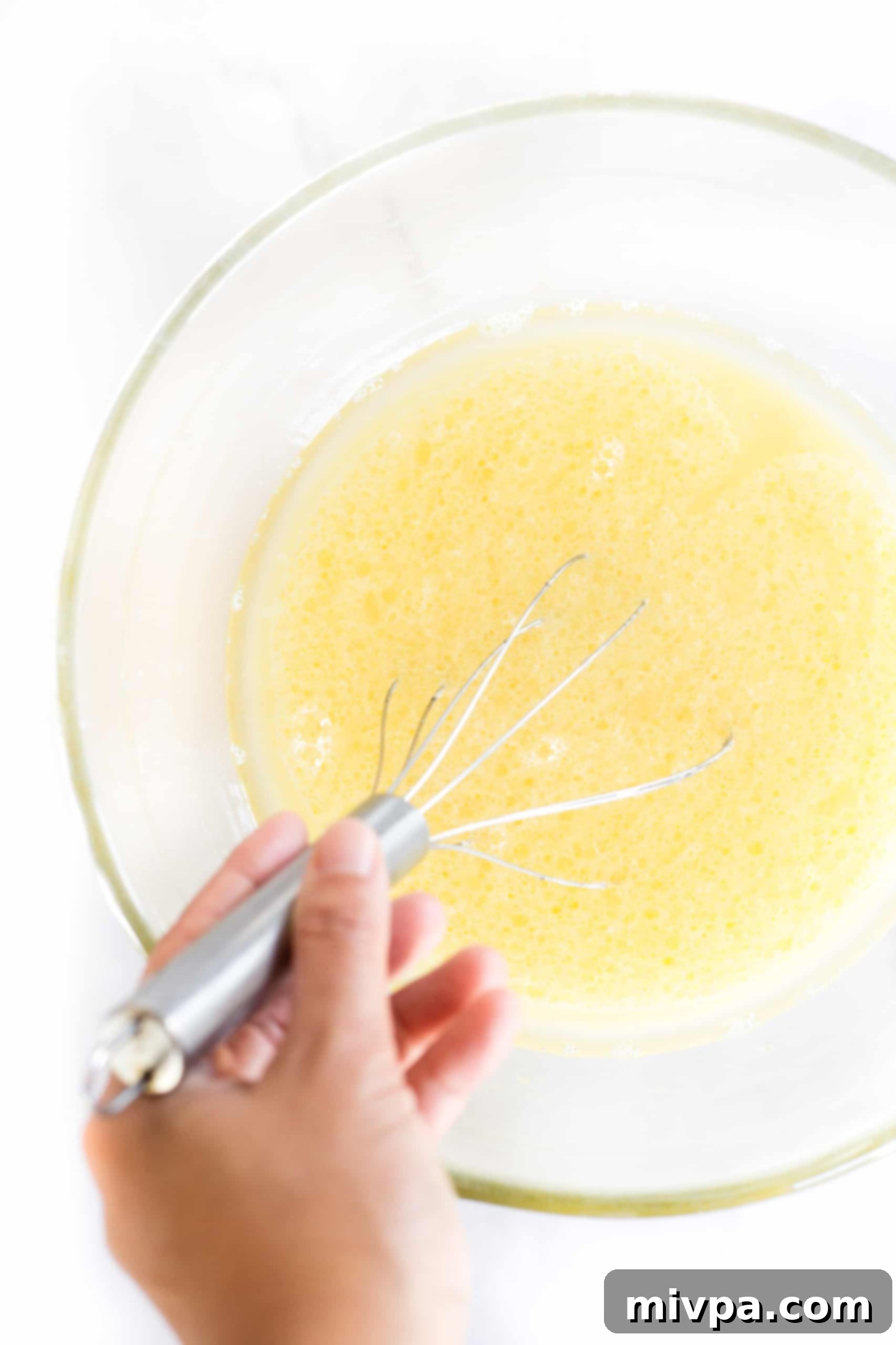Whisking a yellow liquid mixture vigorously in a clear glass mixing bowl.
