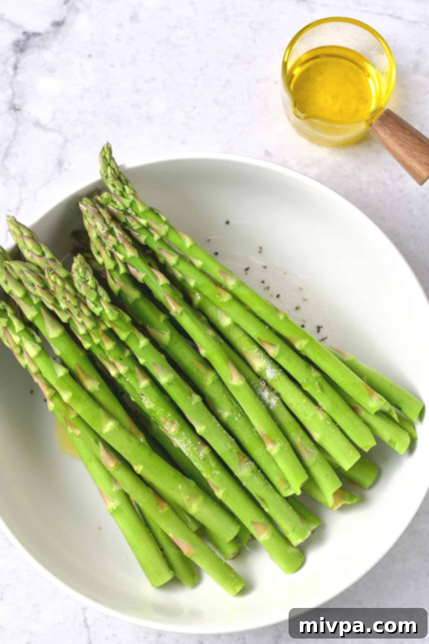 A bowl of fresh asparagus stalks being tossed with olive oil, salt, and pepper for seasoning.