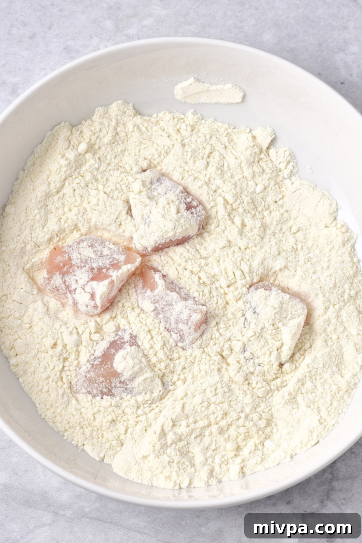 GF Orange Chicken (Dairy-Free) Close-up of chicken pieces being coated in a dry flour mixture within a bowl, ensuring an even layer.