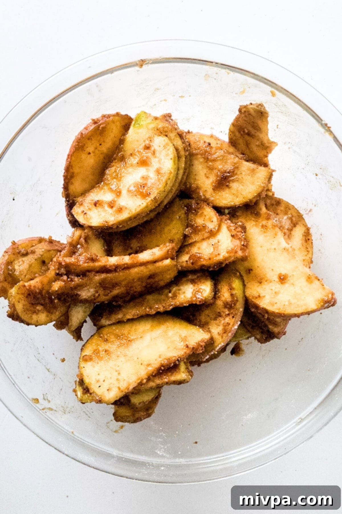 Spice-coated apple slices resting in a clear glass mixing bowl, ready for the crisp.