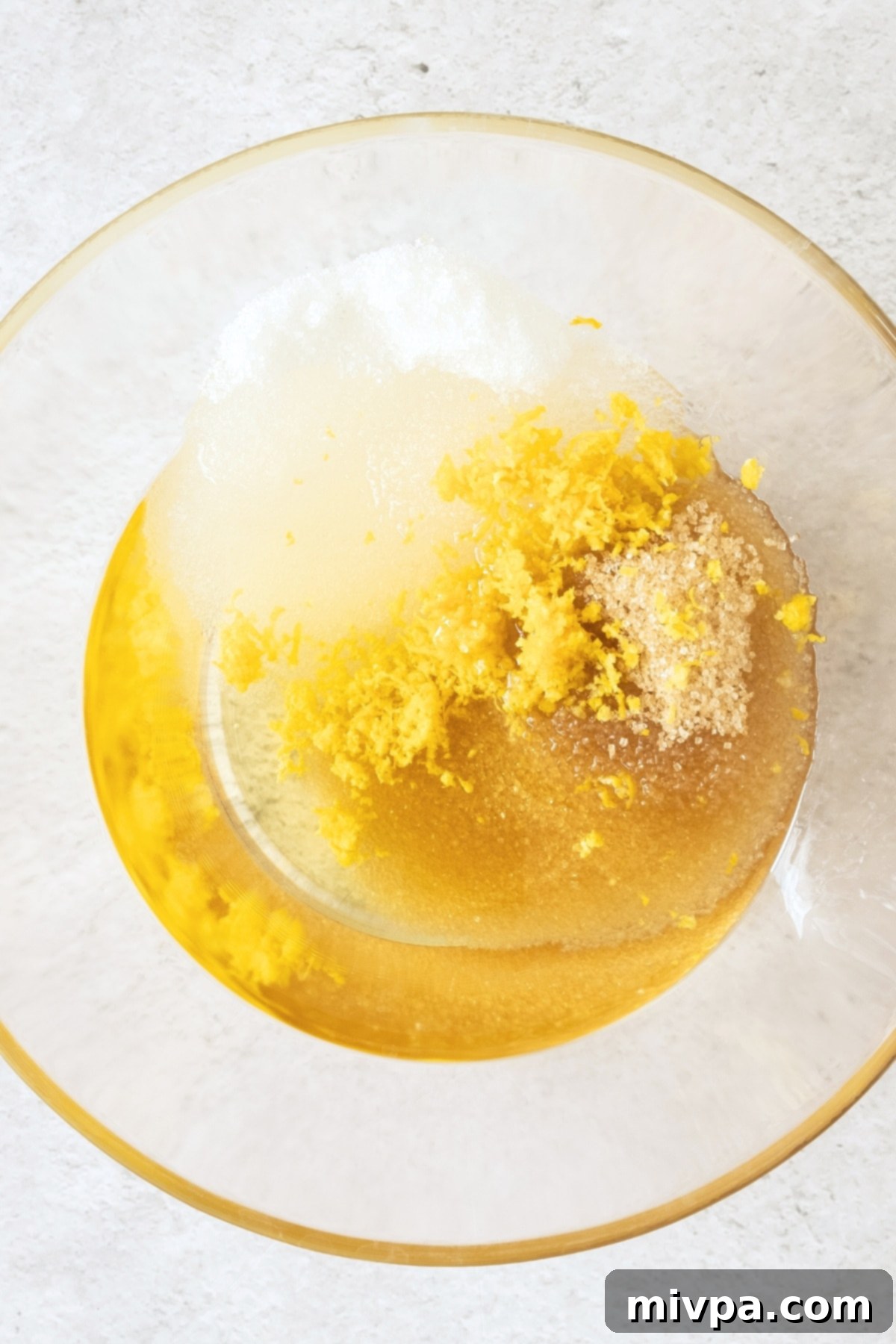 A mixing bowl containing sunflower oil, white sugar, light brown sugar, and lemon zest, ready to be beaten.