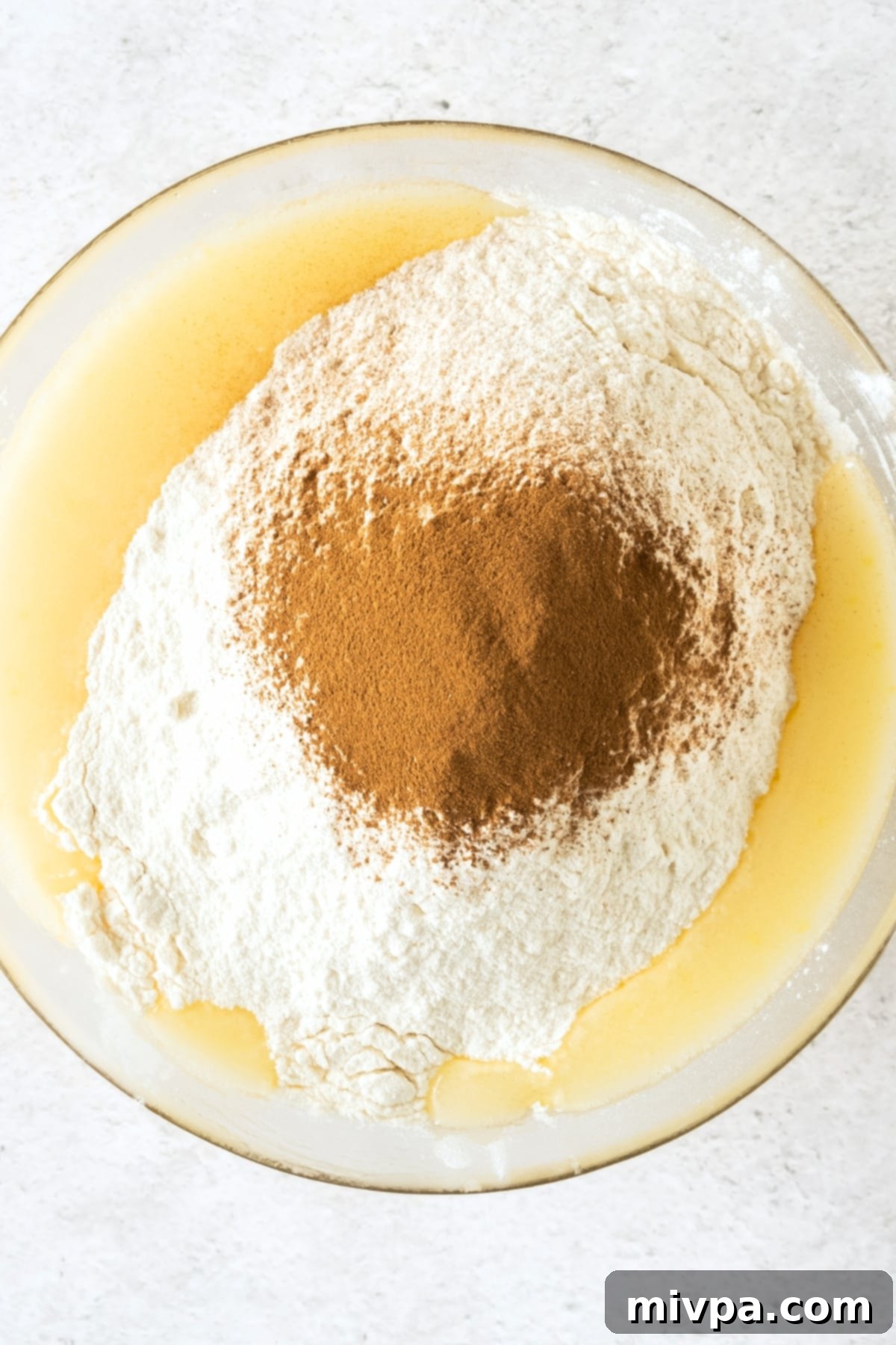 A bowl showcasing the flour mixture and ground cinnamon gently resting atop the pale yellow wet ingredients, ready to be combined.