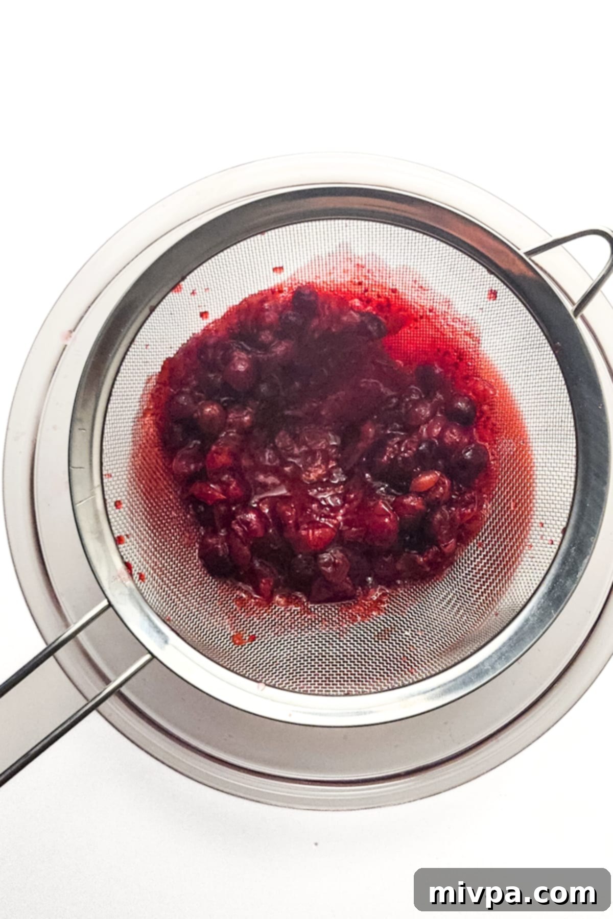 A person using a fine mesh sieve to strain cooked cranberry pulp from the rich red juice into a bowl.