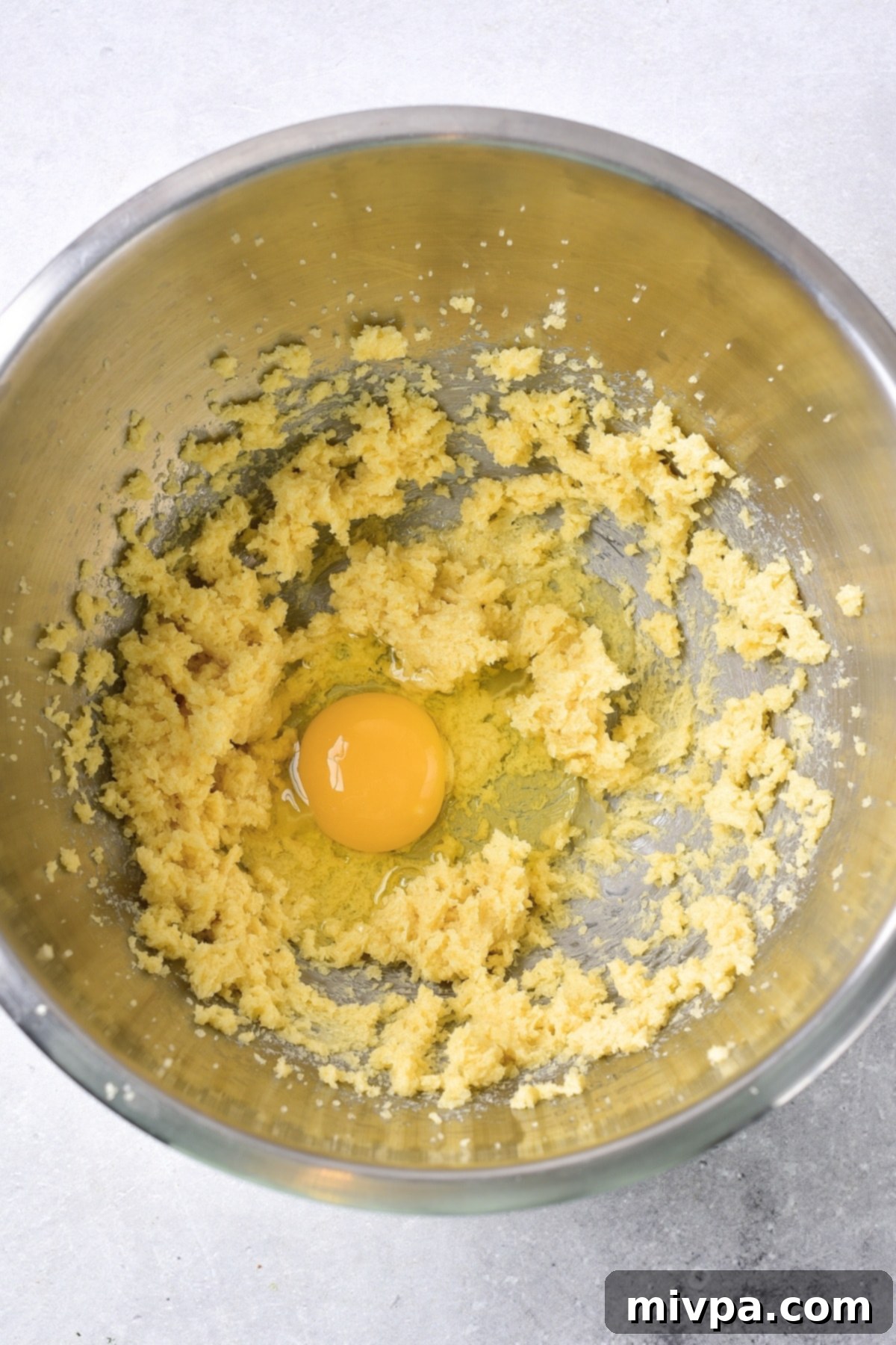 Gluten-Free Dairy-Free Cranberry Bliss Bars (Egg Addition) A single egg cracking into a pale yellow, creamy butter and sugar mixture in a large mixing bowl, mid-process.