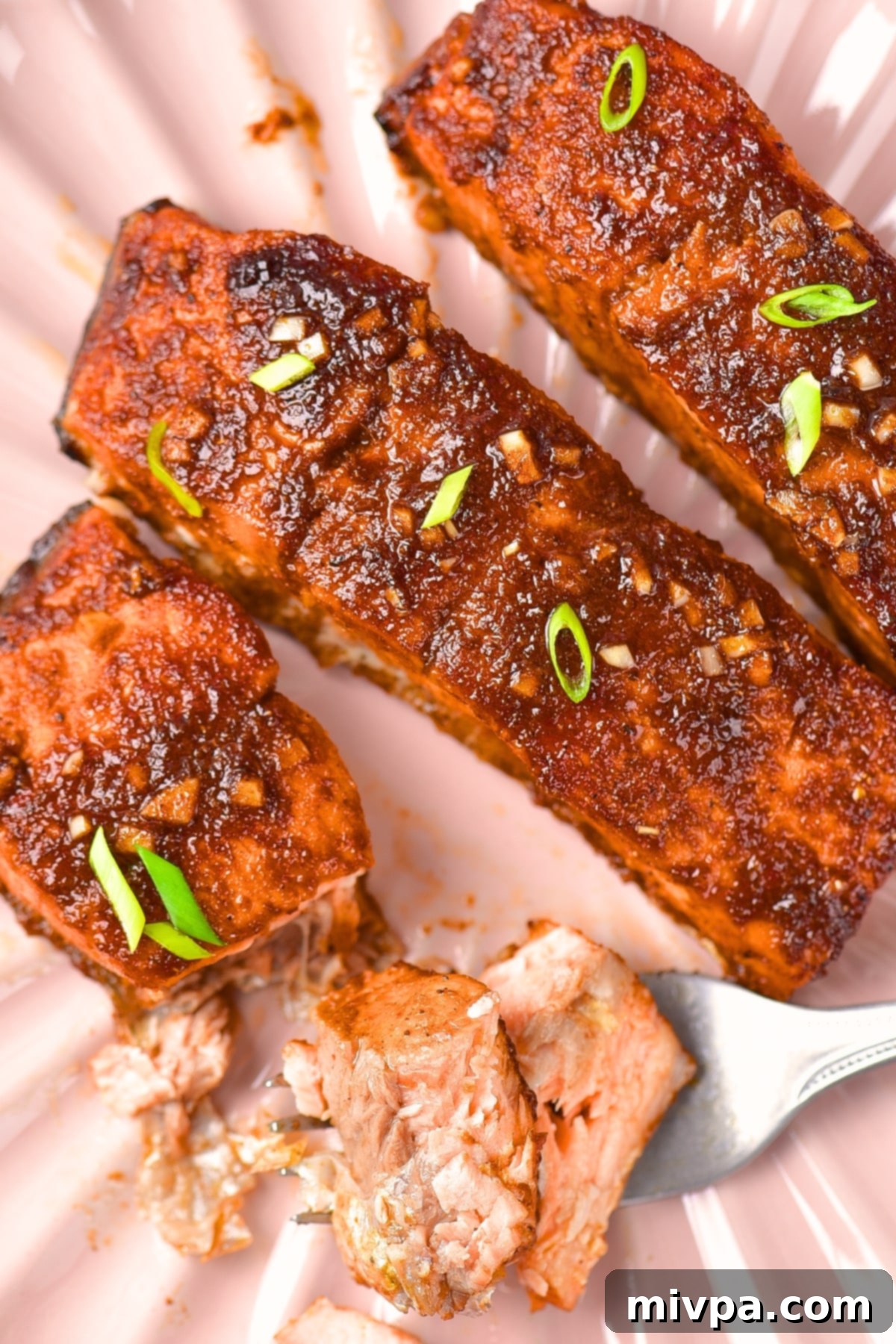 A top-down view of three perfectly baked spicy firecracker salmon fillets on a pink plate, garnished and ready to serve.
