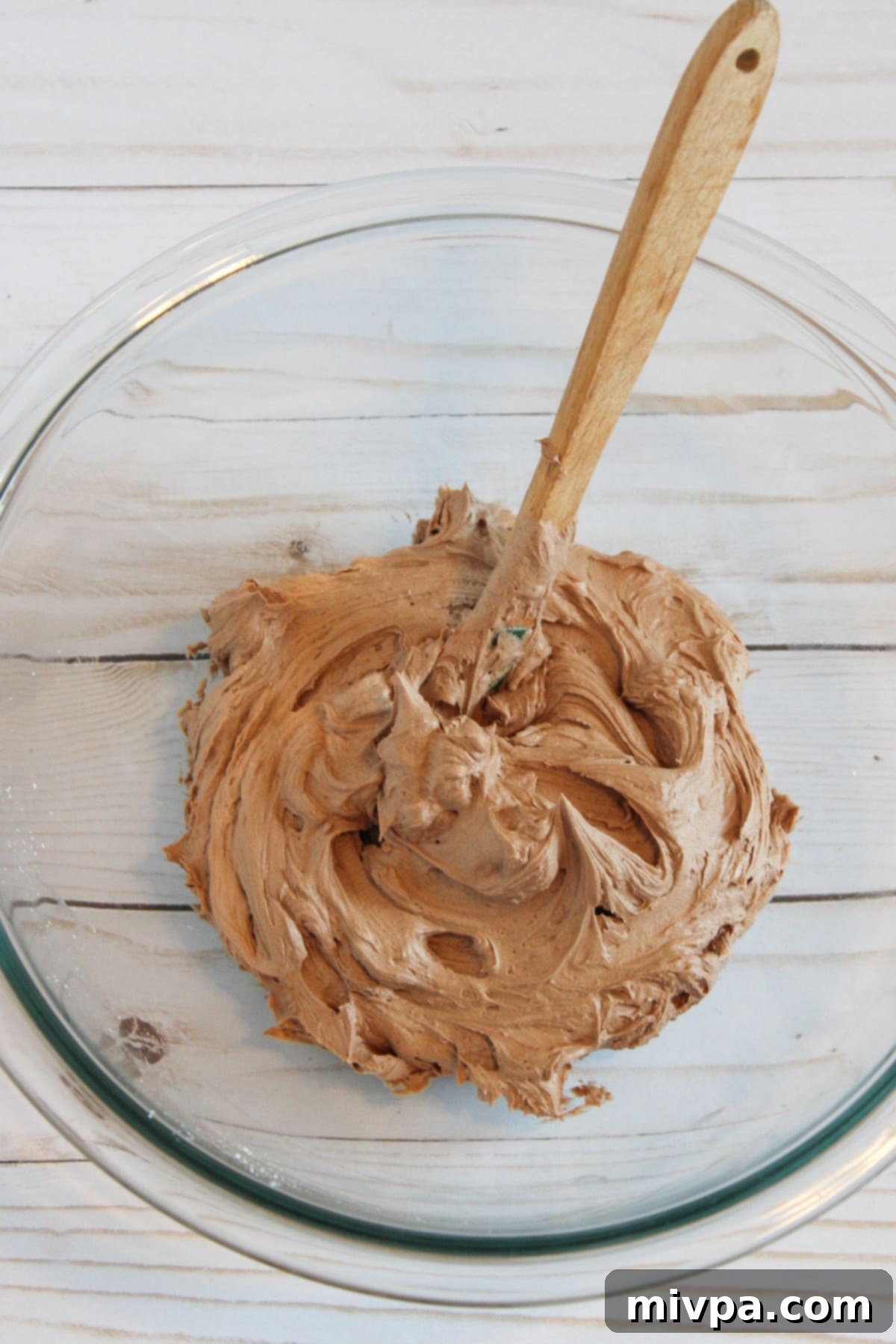 Mixing chocolate buttercream frosting in a clear glass bowl with an electric hand mixer, showing the creamy texture.