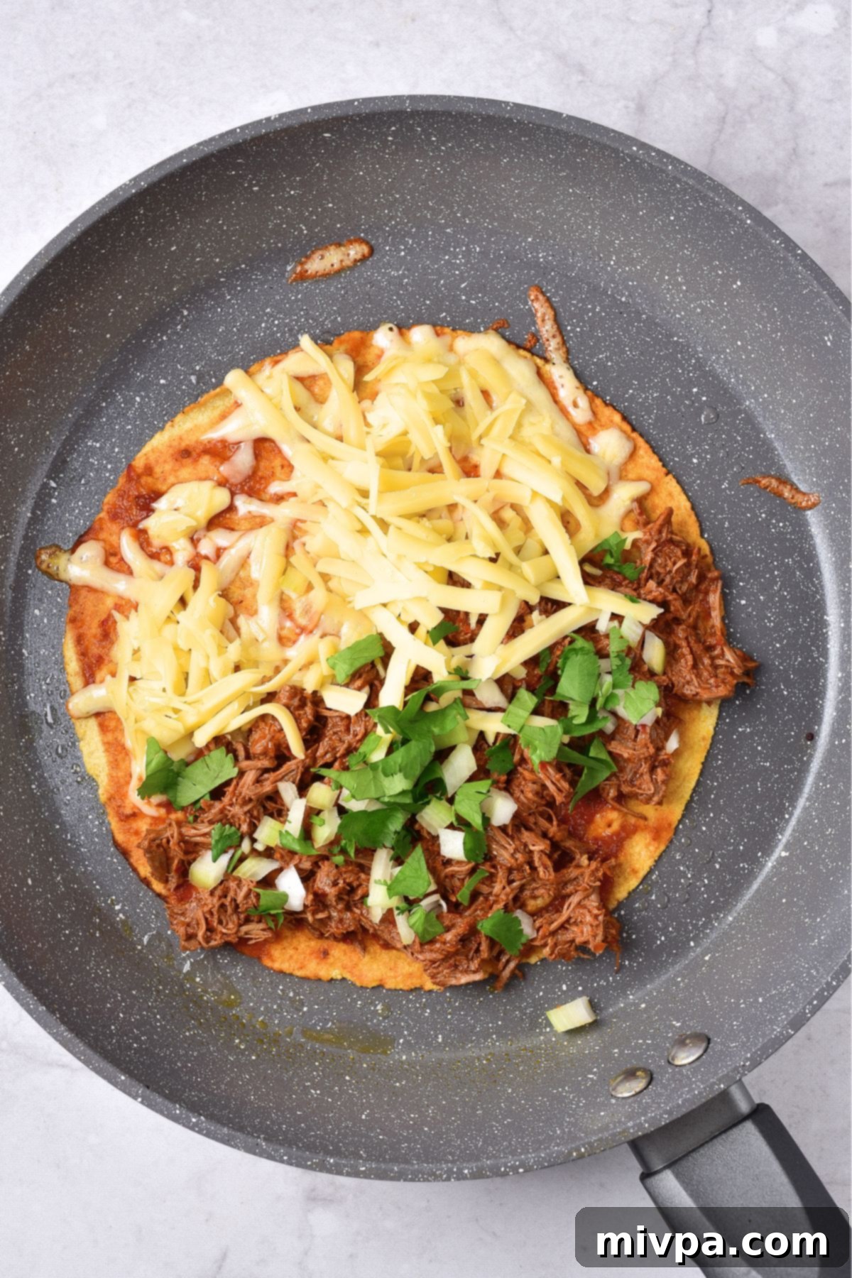 Cooking a tortilla topped with shredded birria beef and shredded cheese in a skillet.