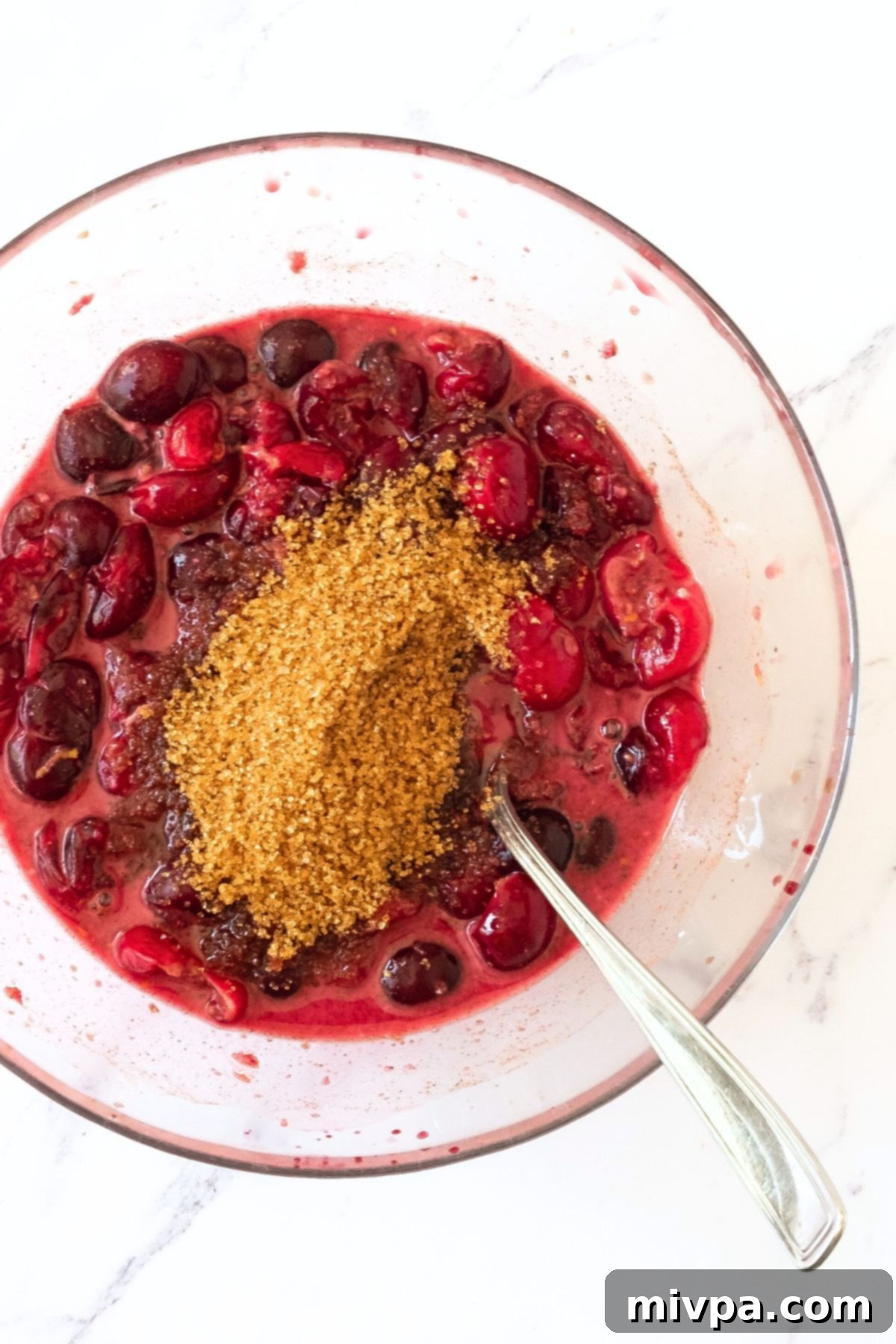 Easy Cherry Crumble (Gluten-Free, Vegan) A clear glass bowl filled with fresh, glistening cherries, topped with brown sugar and spices, ready for mixing to form the crumble filling.
