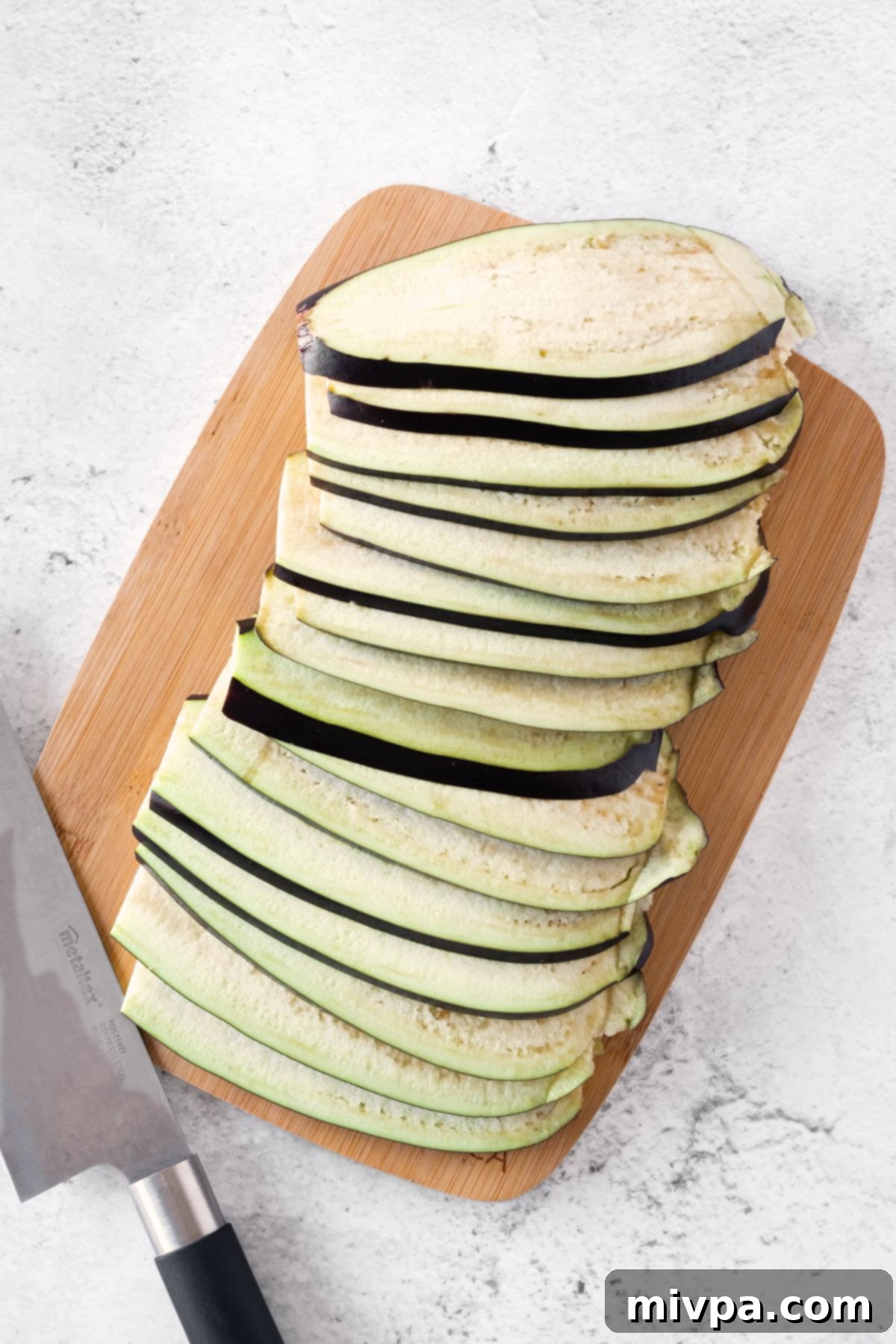 Thinly sliced eggplant pieces neatly arranged on a wooden cutting board, ready for the salting process.