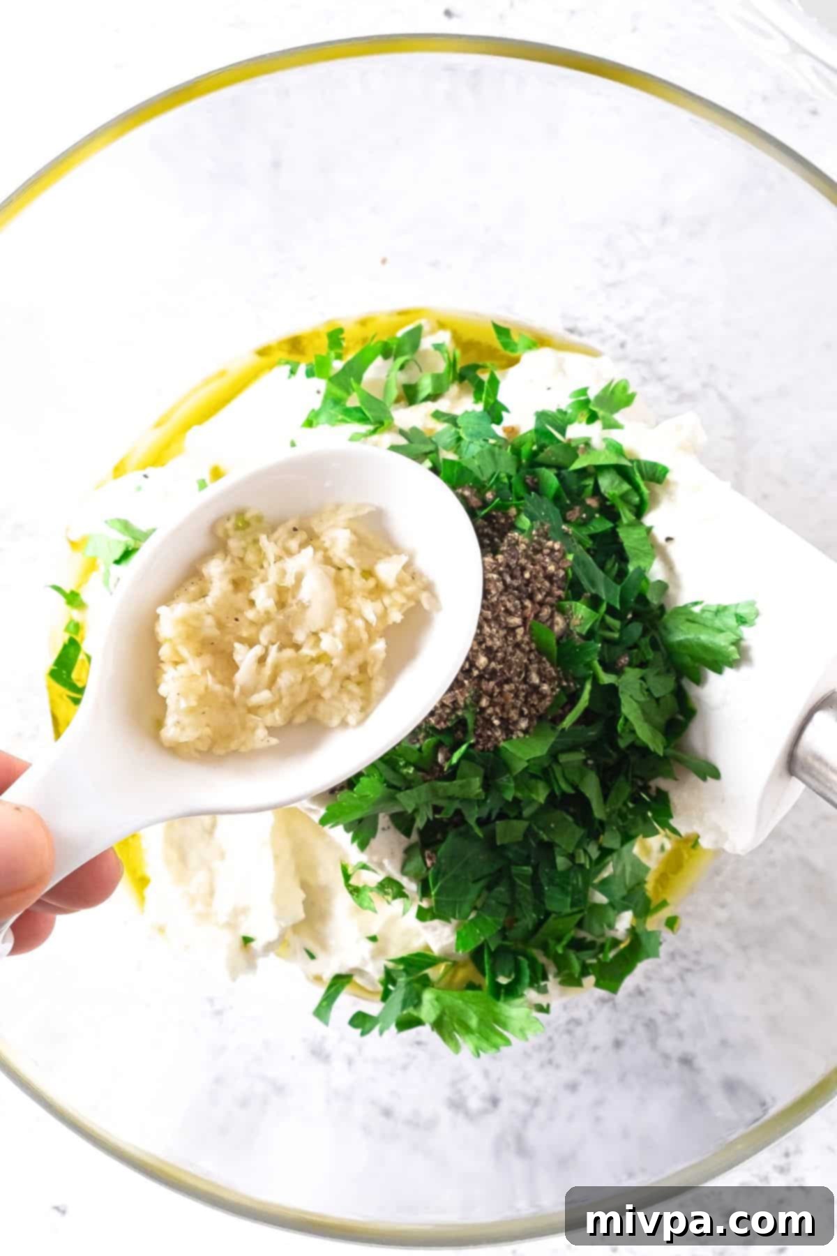 A hand holding a spoonful of grated garlic, ready to be added to a bowl containing cream cheese, Greek yogurt, parsley, and other filling ingredients.