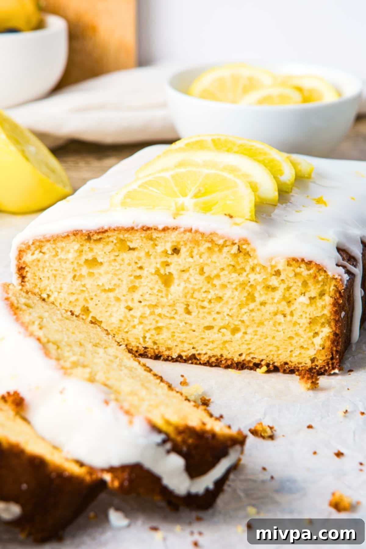 Moist Gluten-Free Lemon Drizzle Cake (Dairy-Free) Half-sliced loaf of gluten-free lemon drizzle cake on parchment paper, showcasing its moist interior and glistening drizzle.