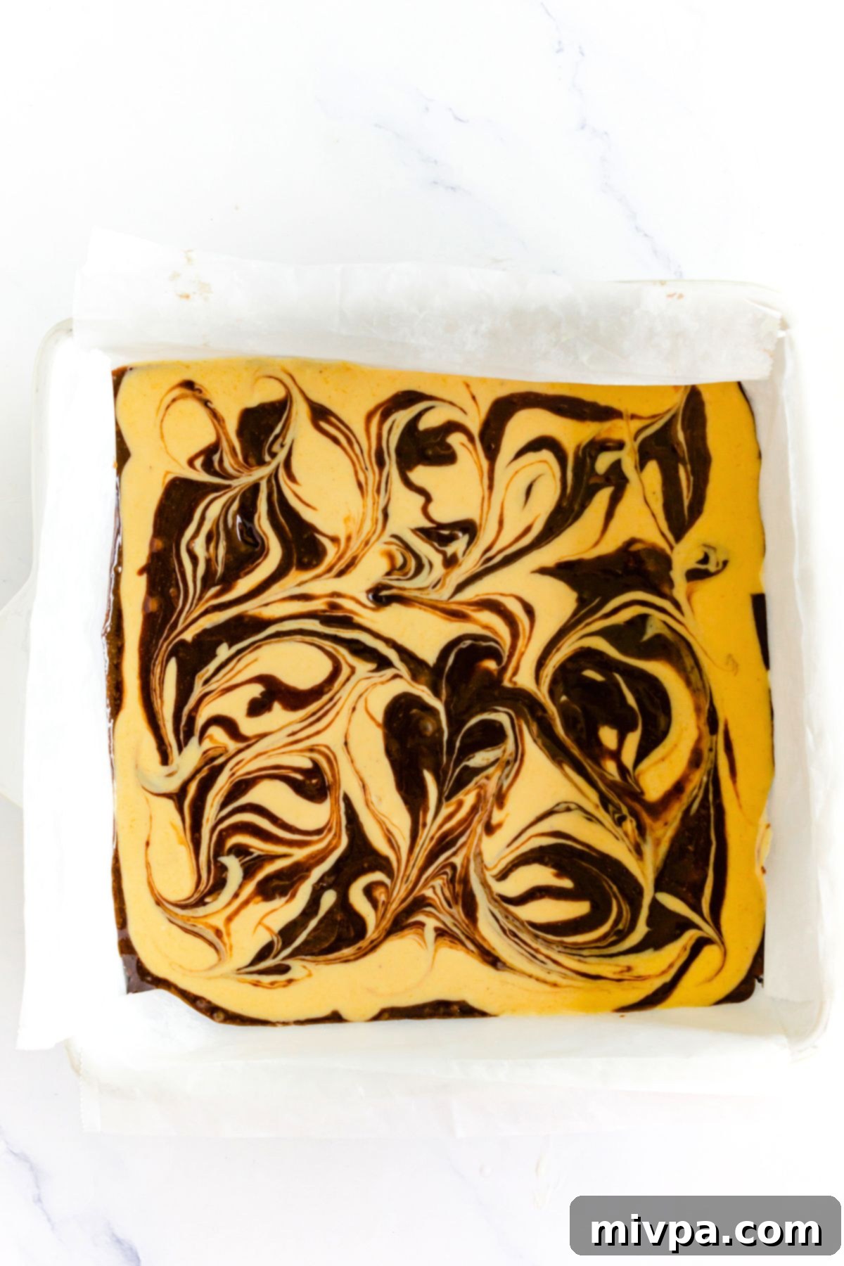 Gluten-Free Pumpkin Cheesecake Brownies Swirl Beautifully swirled brownie and pumpkin cheesecake batter in a parchment-lined baking pan, ready for baking.
