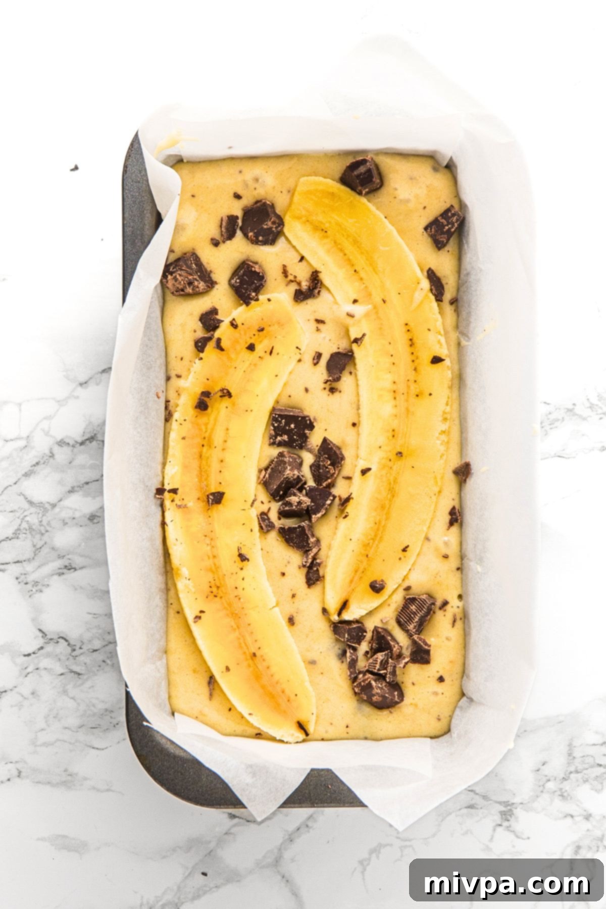 Sliced banana and chocolate on top of batter in parchment-lined loaf pan.
